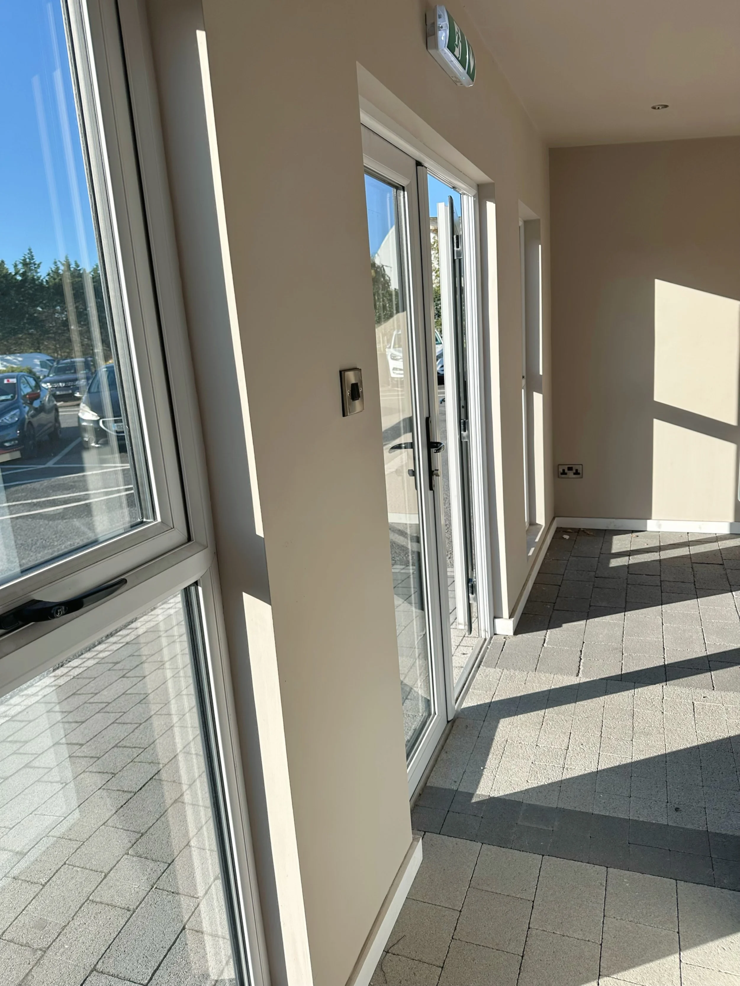 Interior of a building with large windows and a glass door, sunlight casting shadows on the tiled floor, with parking lot visible outside.
