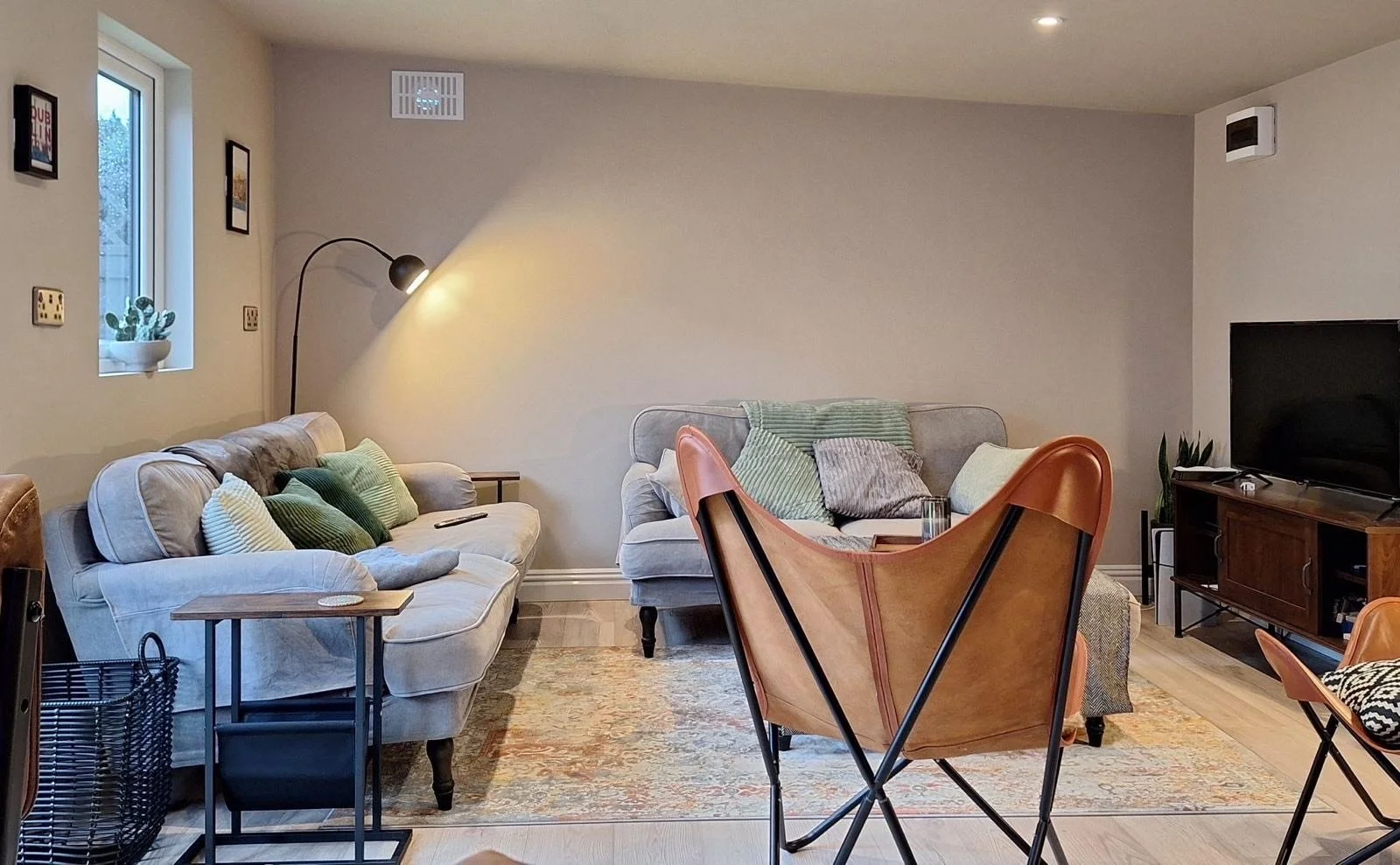 Living room with two beige sofas with green and white pillows, a wooden side table, a black floor lamp, a patterned area rug, a wooden TV stand with a large flat-screen TV, leather and fabric chairs, a window with a small potted cactus, and a beige w