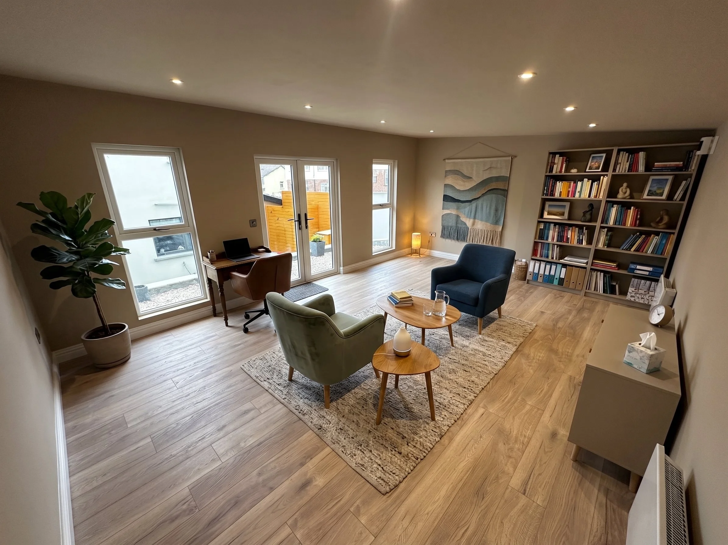 Living room with beige walls, large windows, and wooden flooring, furnished with a green armchair, a blue armchair, a wooden coffee table, a tall bookshelf filled with books, and a small desk with a chair. A large wall hanging and a floor lamp are al