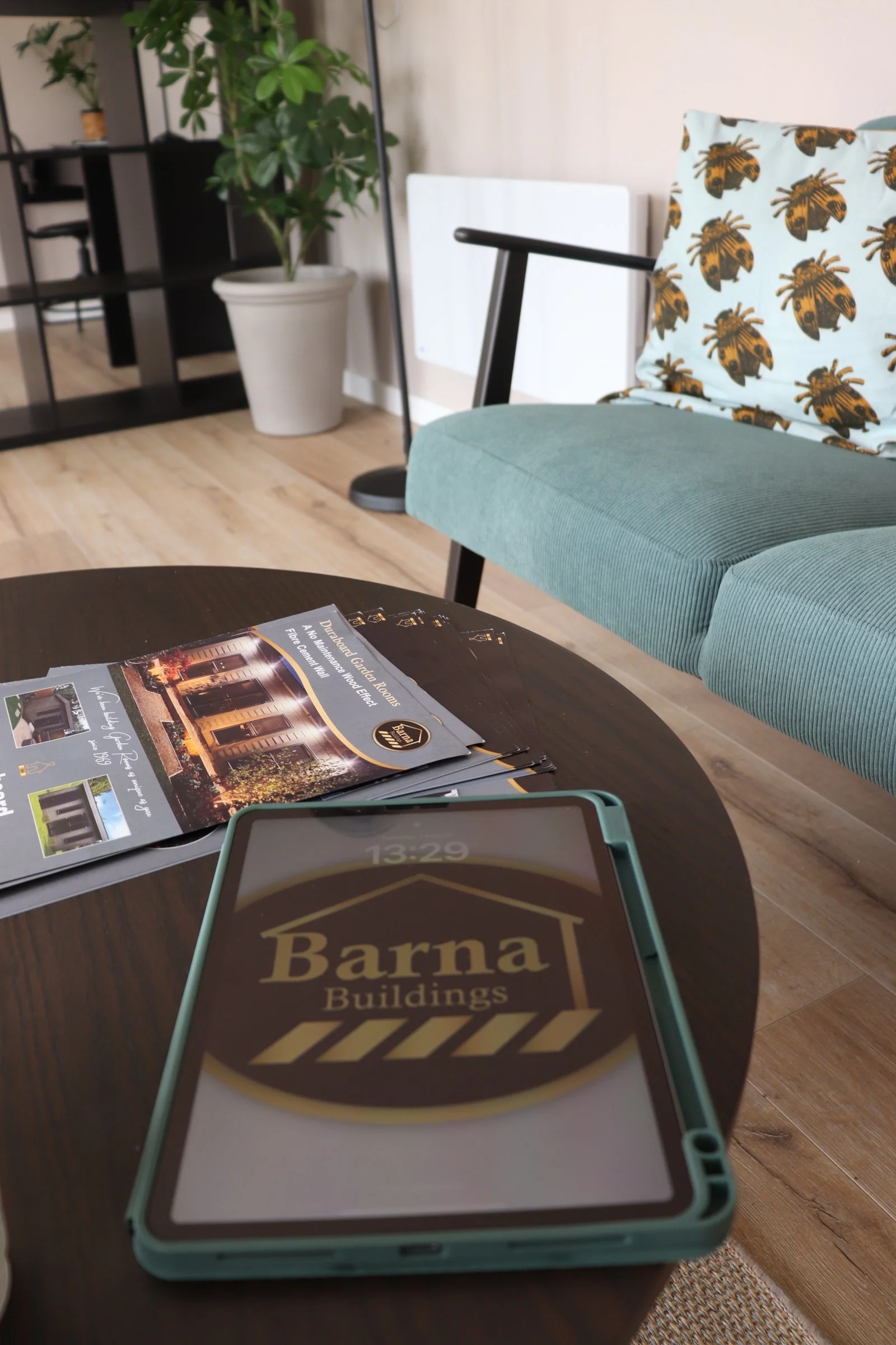 A tablet on a wooden table displaying the Barna Buildings logo, with brochures and a teal cushioned chair with a bee-patterned pillow in the background inside a modern living room or office space.
