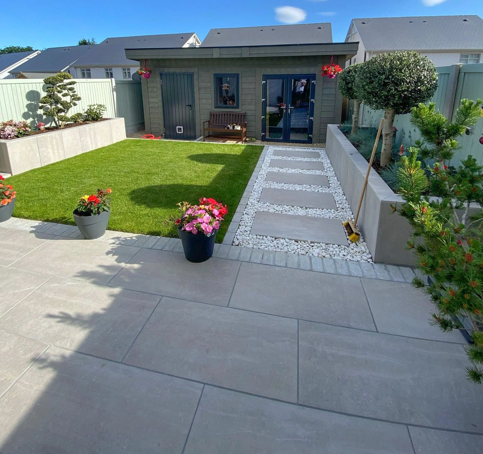 A backyard garden with a concrete patio, potted flowers, a grassy area, and a pathway made of stepping stones with white rocks on either side. There is a small shed in the background, and the area is enclosed by a light green fence. There are trees a