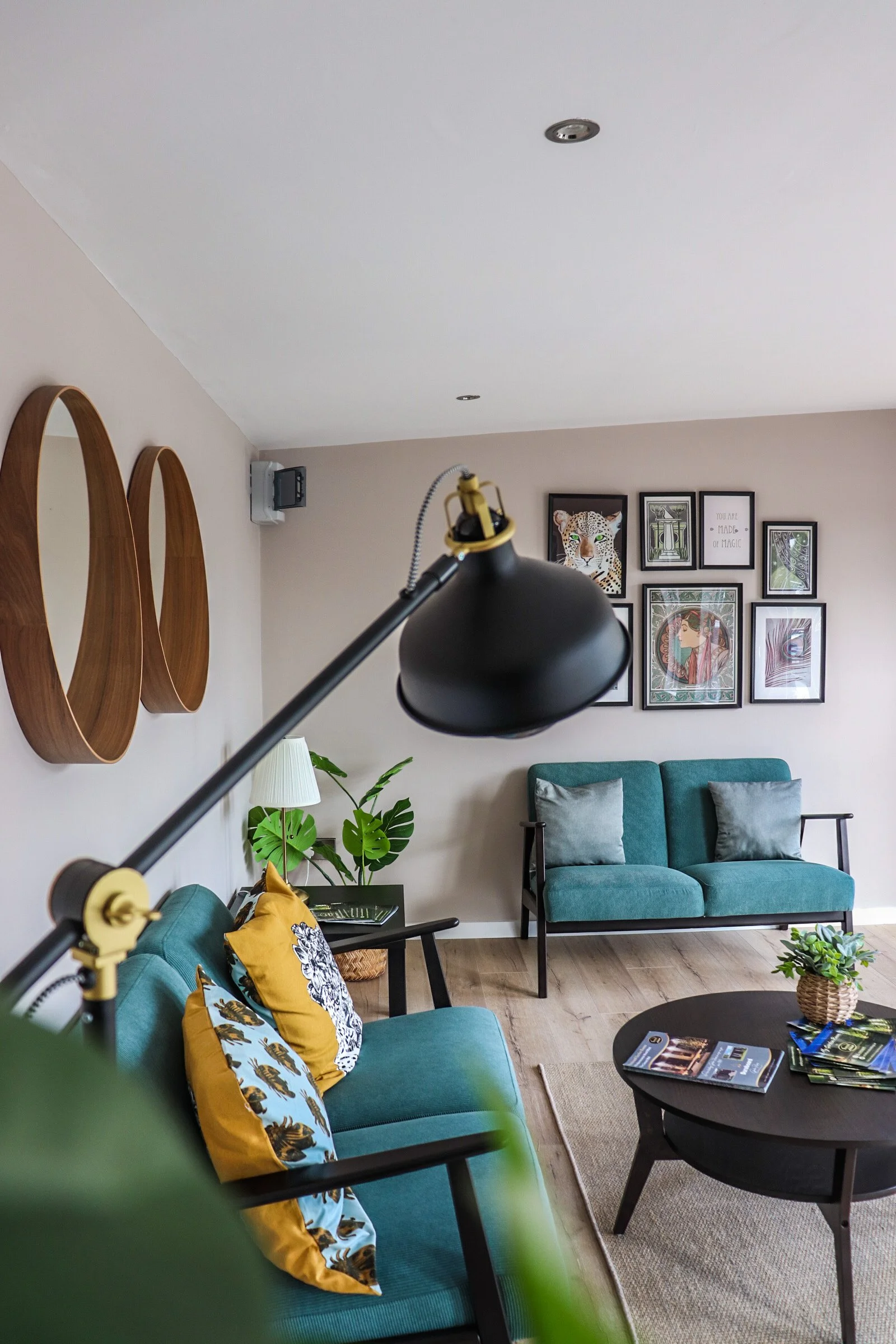 Living room with teal sofas, yellow and patterned pillows, a black coffee table with magazines, framed wall art, a green plant, a floor lamp, and wooden wall decorations.