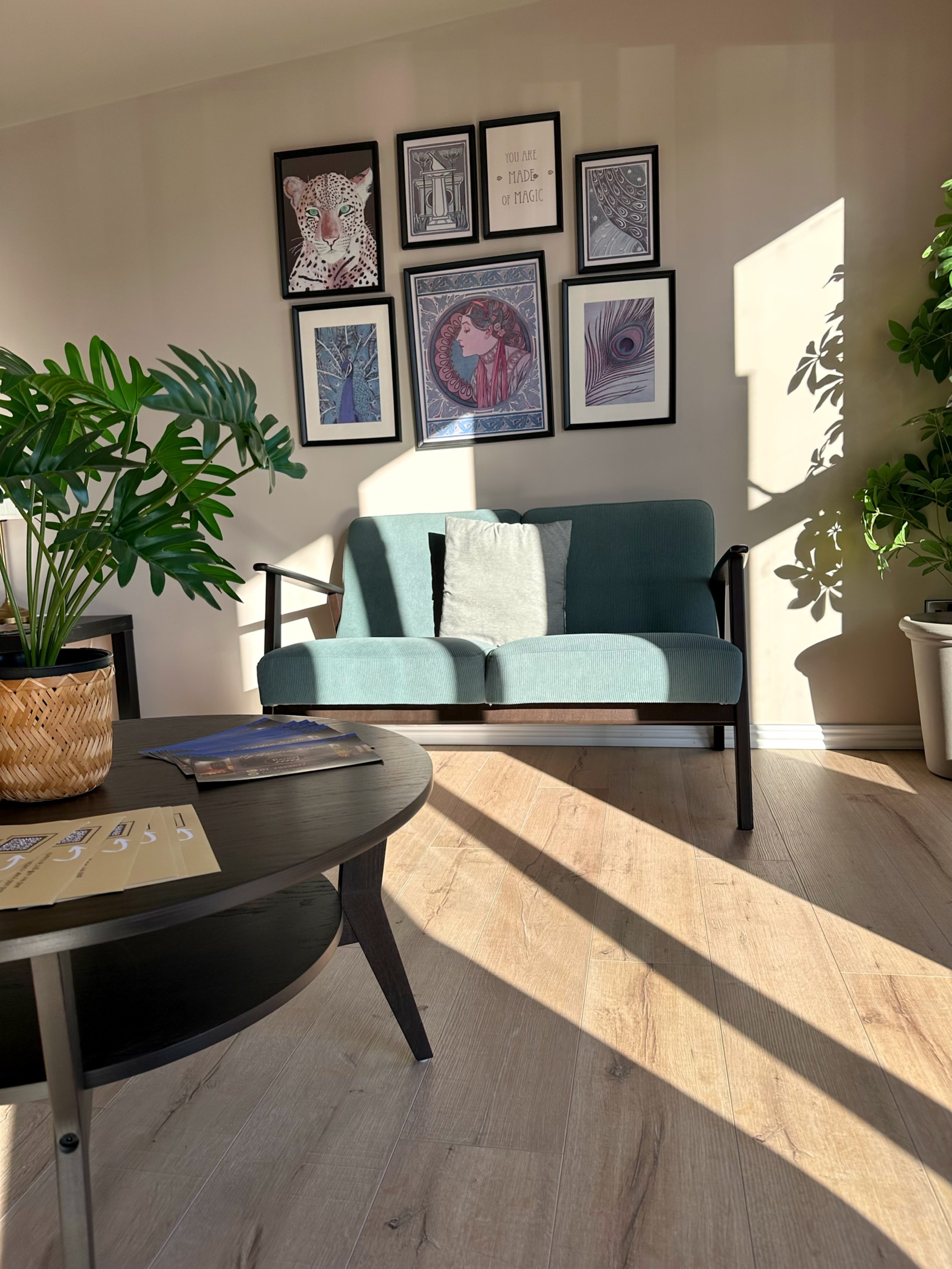 A cozy living room corner with a teal sofa, decorative pillows, a circular dark wood coffee table with brochures, large potted plants, and a wall gallery with seven framed pictures, including an image of a leopard, abstract art, and a motivational qu