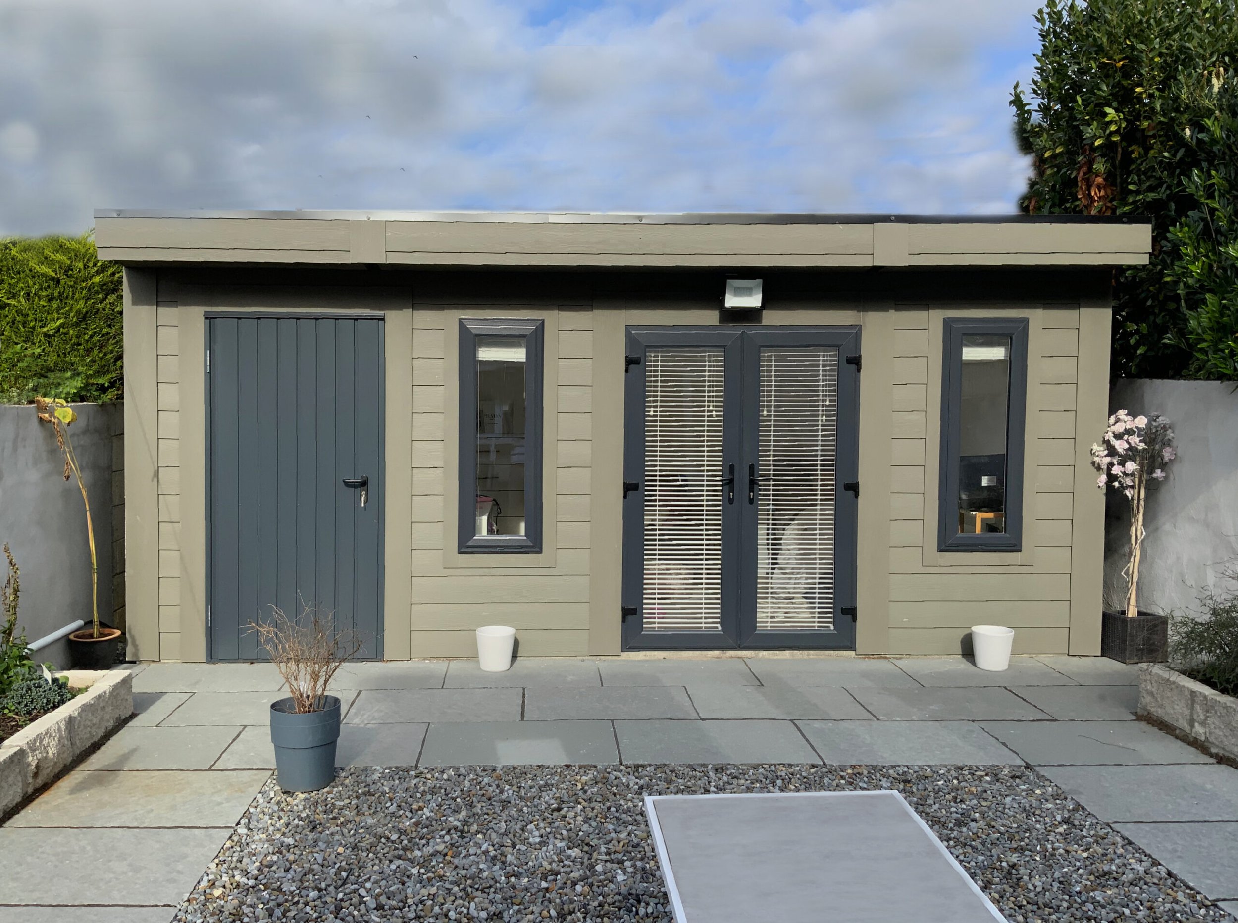 Small gray garden shed with three windows, double doors with blinds, and a small gray door, set in a paved backyard with potted plants and a gravel bed.