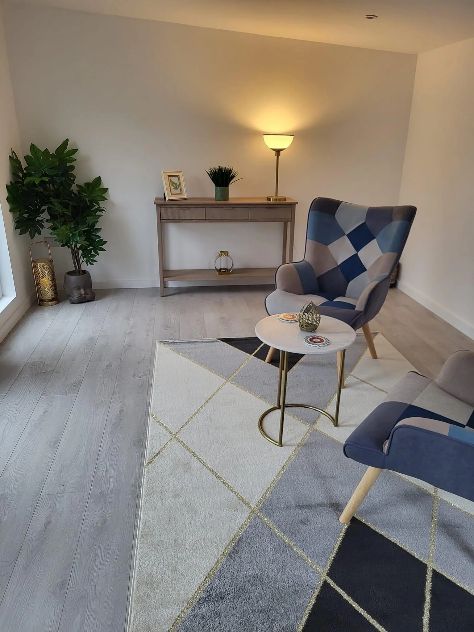 A cozy living room corner with modern decor, including a patterned armchair, a small round coffee table, a side table, a floor lamp, and decorative plants on a light-colored rug.