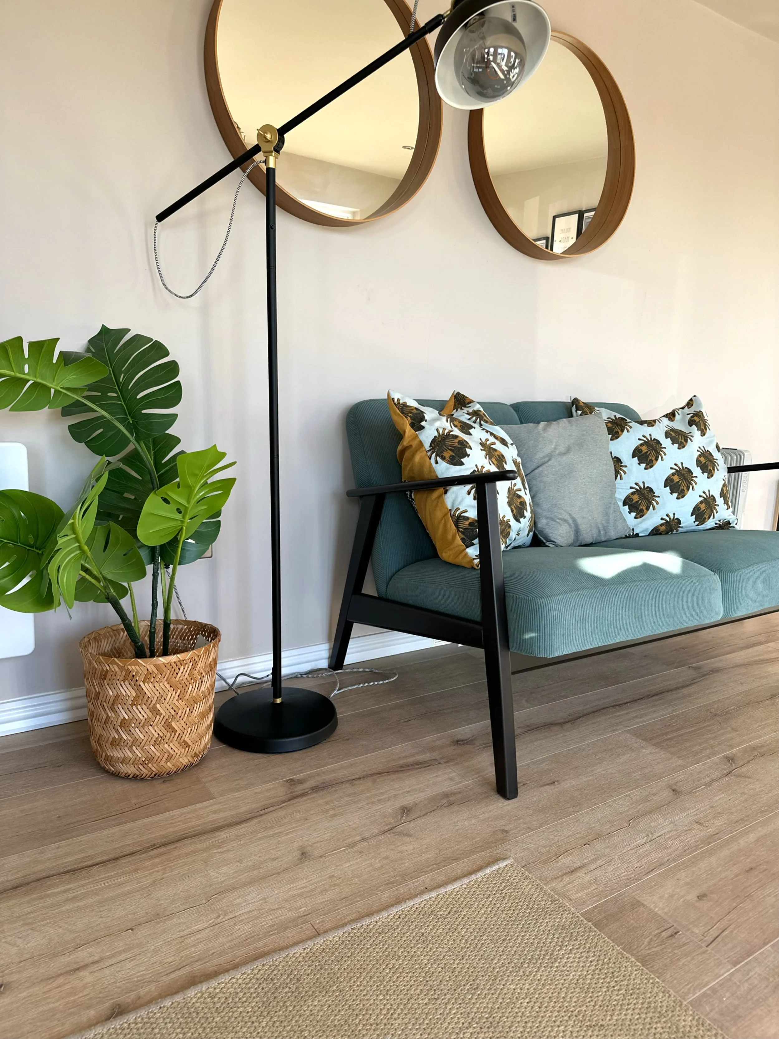 Living room with a green sofa, decorative pillows with tropical leaf patterns, a large leafy green plant in a woven basket, a modern black and gold floor lamp, two round wooden mirrors on the wall, and wooden flooring.