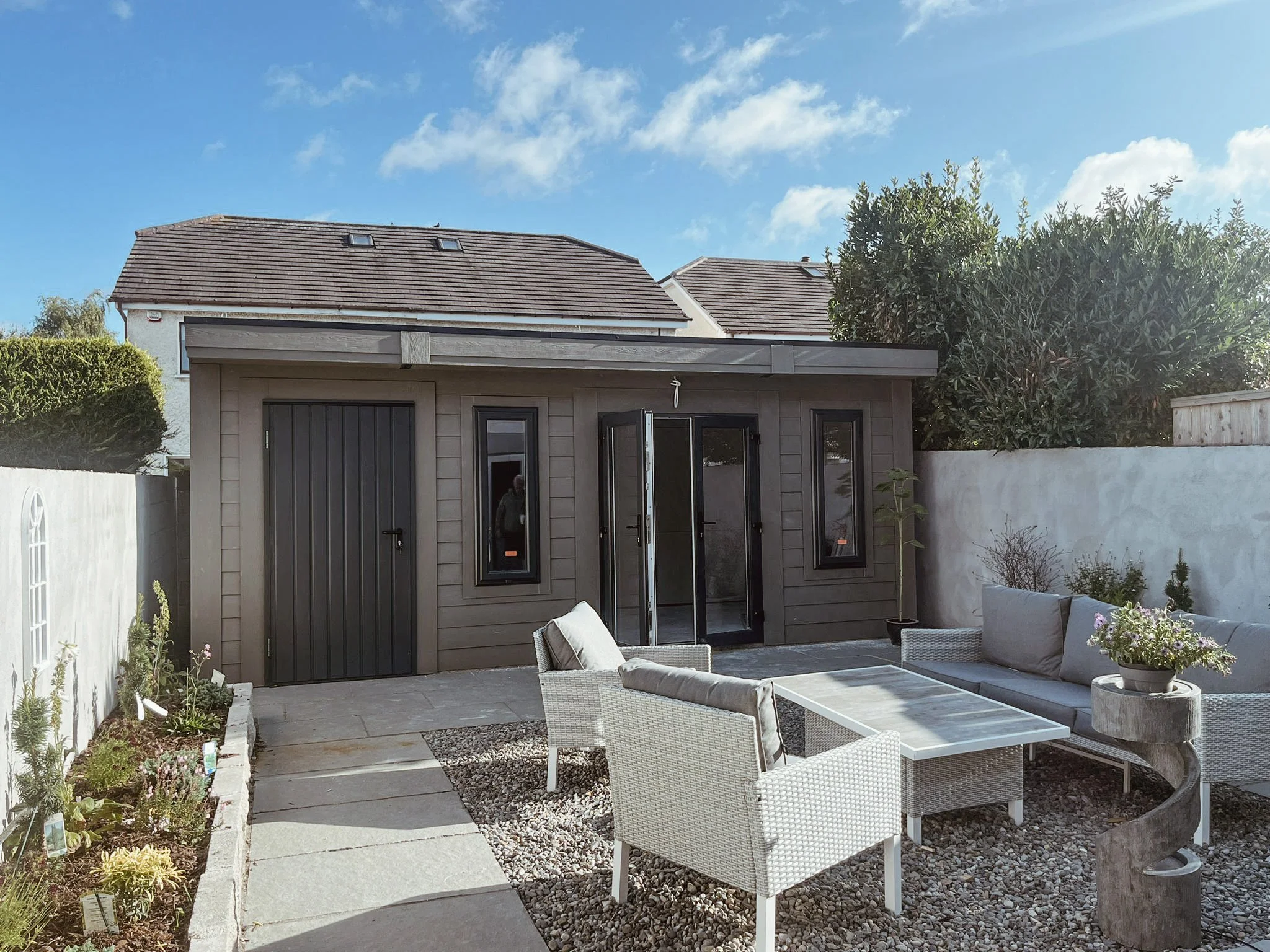 Backyard patio with outdoor seating, gray and white furniture, a small building with black doors and windows, concrete pathway, gravel ground, garden plants, and a tall green hedge with a large tree in the background under a partly cloudy sky.
