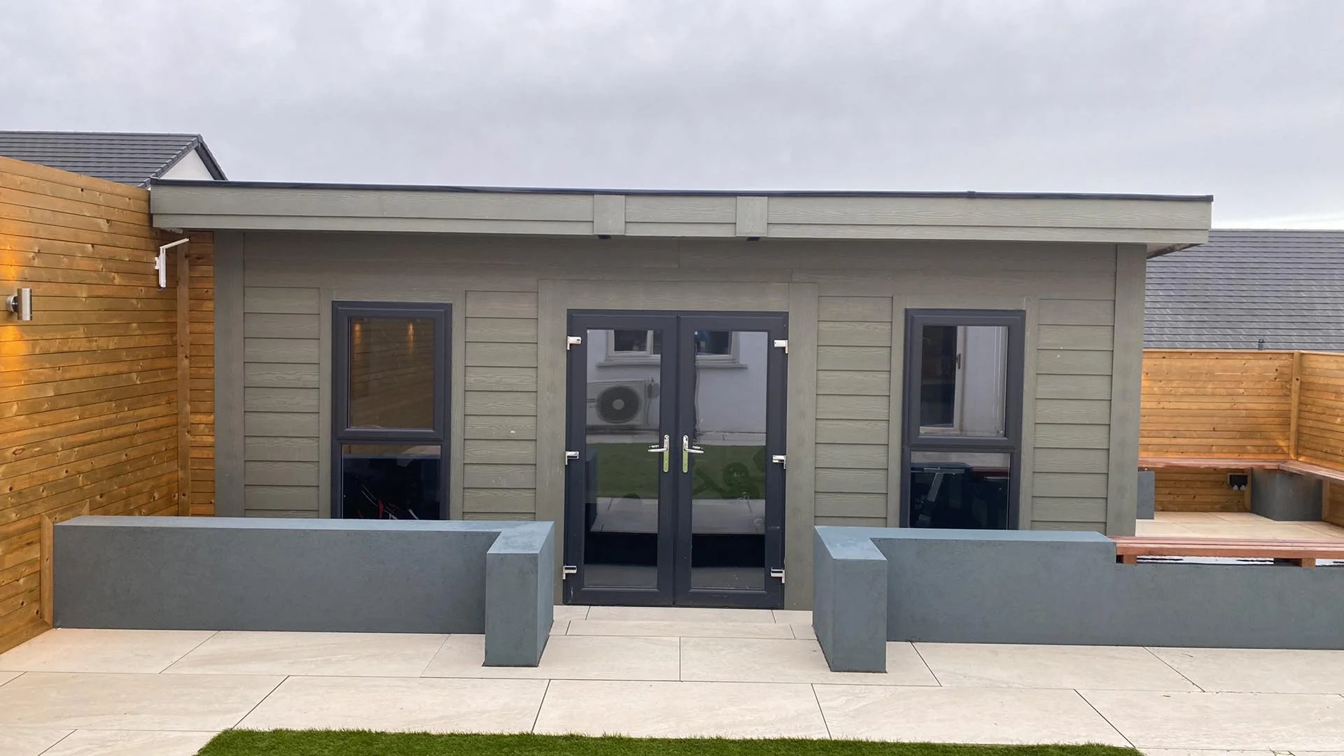 Modern backyard shed with gray wood siding, black-framed glass double doors, and matching windows on either side. The shed is enclosed by wooden fencing with a built-in bench on the right side. There are concrete planters in front and a paved patio a