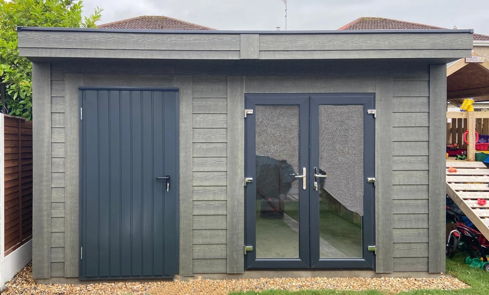 Gray wooden shed with a single-pane door on the left and double-pane door with mesh on the right, located in a backyard with a gravel ground.