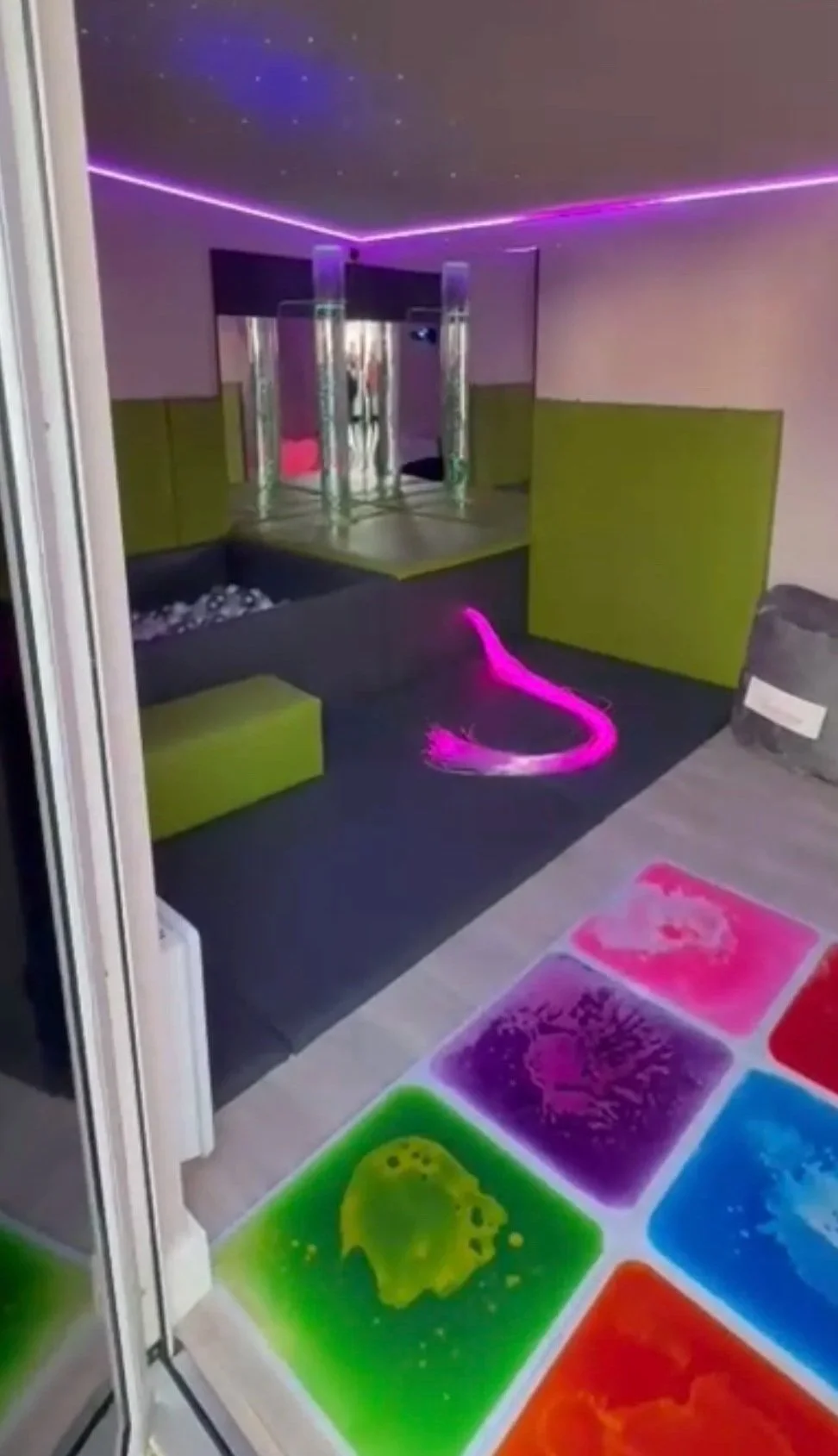 Colorful glow-in-the-dark tiles on the floor, a small sun-shaped decoration, modern living room with neon lighting, and a mirrored wall.