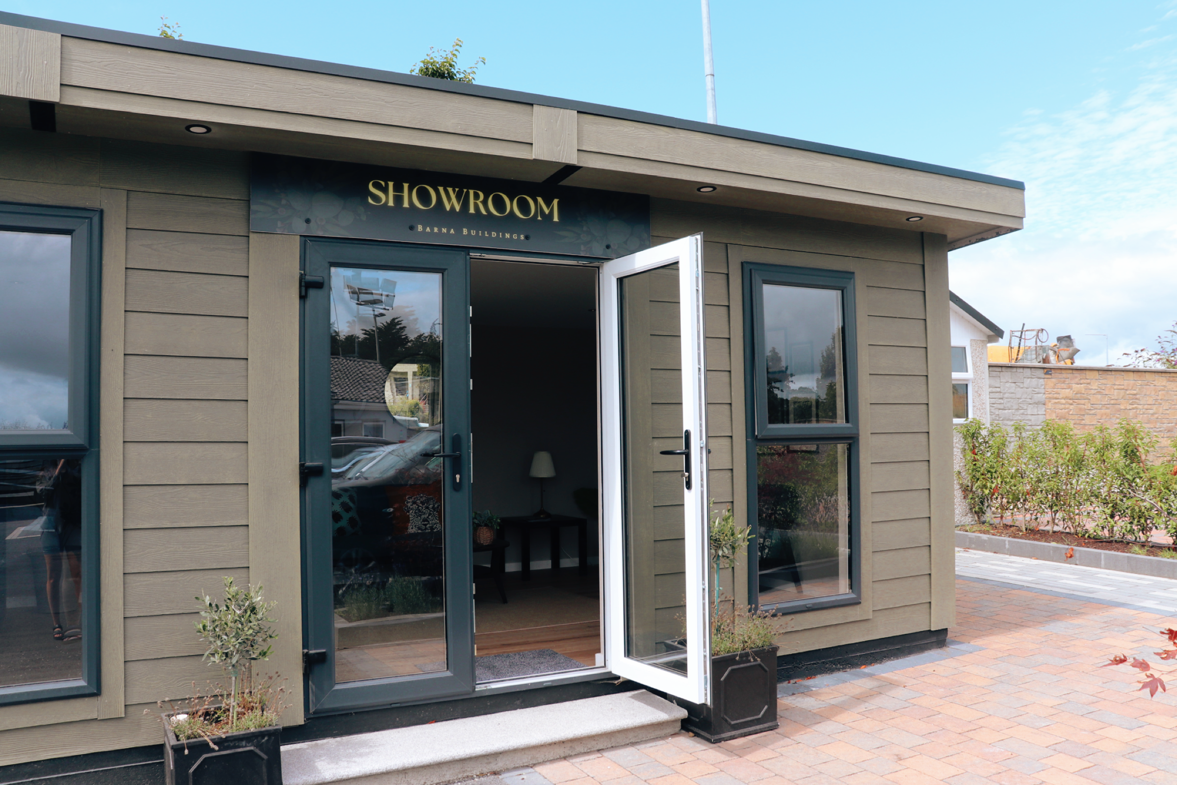The exterior of a building with a sign that says 'Showroom' and open glass doors, with potted plants outside and a paved walkway.