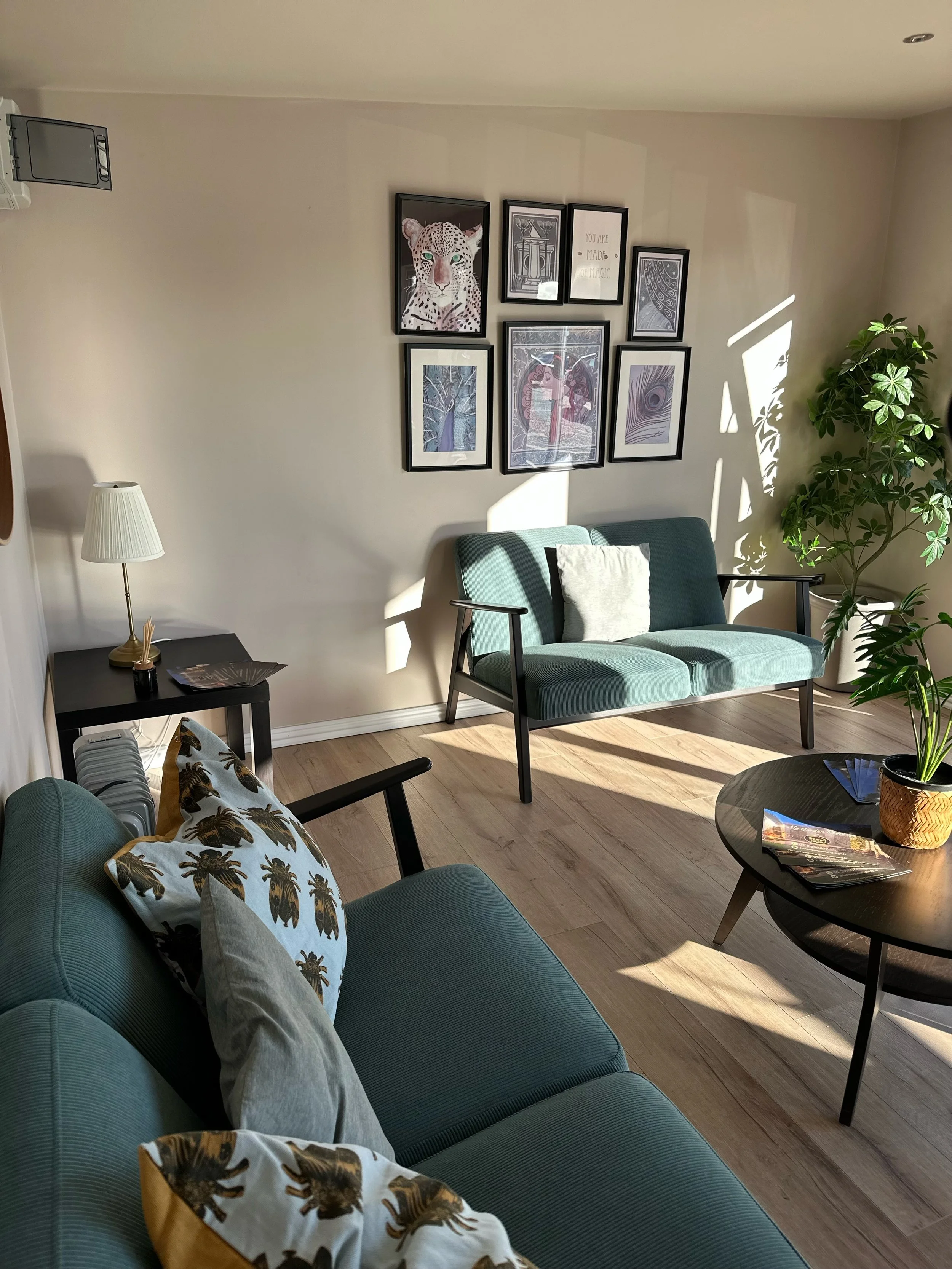 Living room with green sofas, a black coffee table with magazines, a lamp on a side table, framed artwork on the wall, and a large potted plant, with sunlight casting shadows.