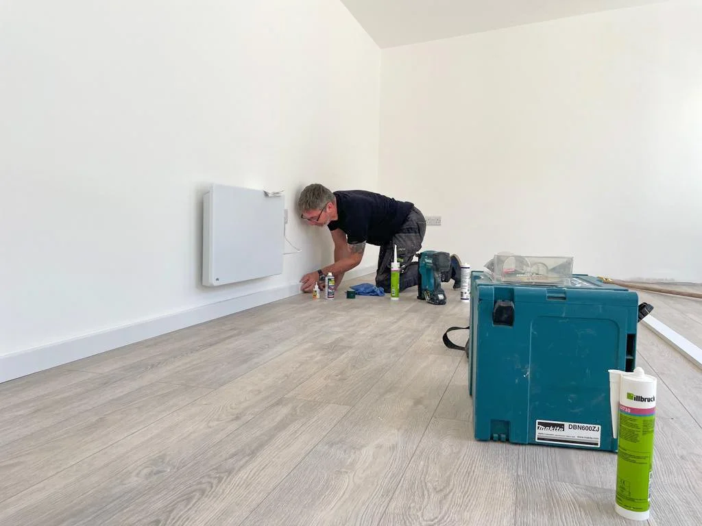 A man installing or repairing a wall-mounted heater in a room with light-colored wood flooring and white walls. Tools and supplies are spread around him. A+ Rated laminate flooring and Farrow and Ball paint.