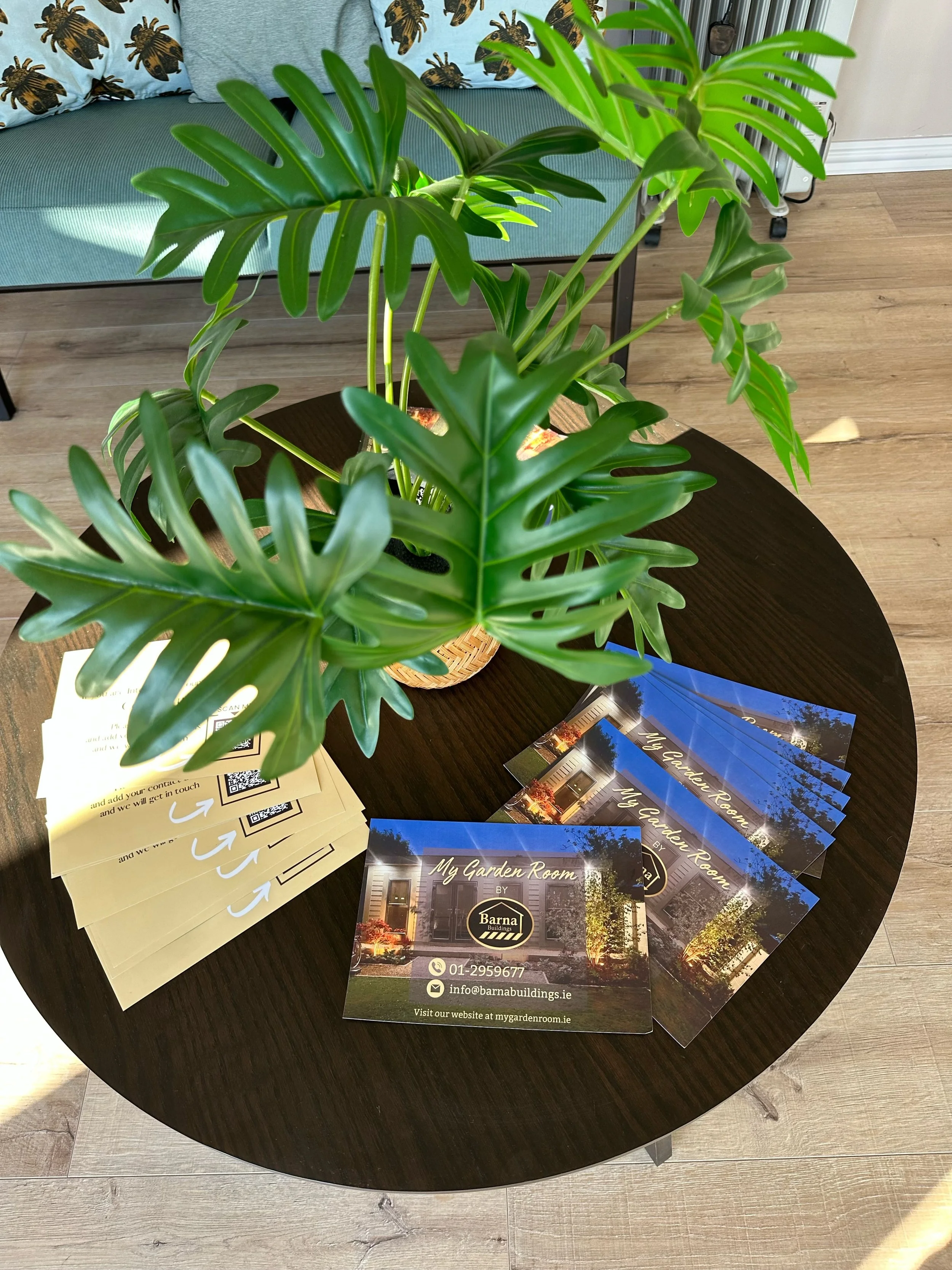 A dark wooden round table with a large green monstera plant in a woven basket, several yellow flyers with a QR code and contact information, and multiple brochures titled "My Garden Room" by Barnat Buildings, with a house and garden scene on the cove