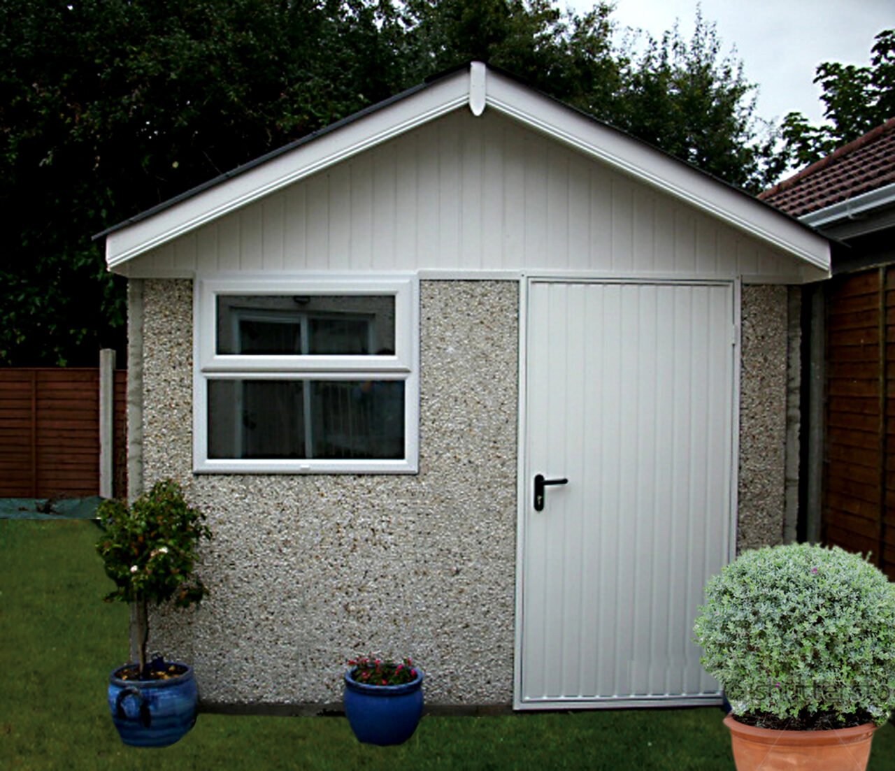 Small garden shed with textured exterior, white door and window, surrounded by potted plants and grass, with trees and neighboring roof in background.