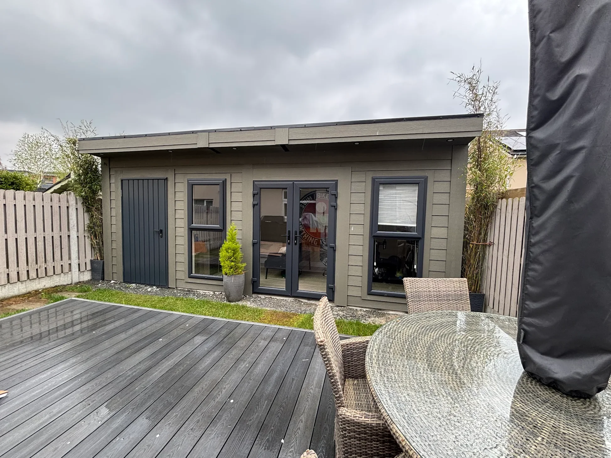 A backyard patio with a gray wooden deck, outdoor dining table with chairs, and a small gray building with black-framed doors and windows, under a cloudy sky.
