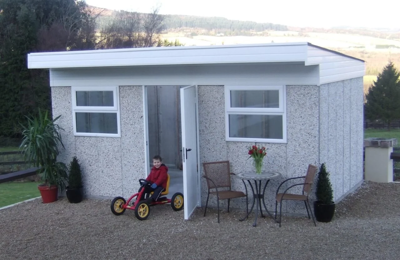 A young boy on a tricycle outside a small, white, textured building with two windows, a table with a vase of flowers, and two chairs, set against a rural landscape with trees and hills in the background.