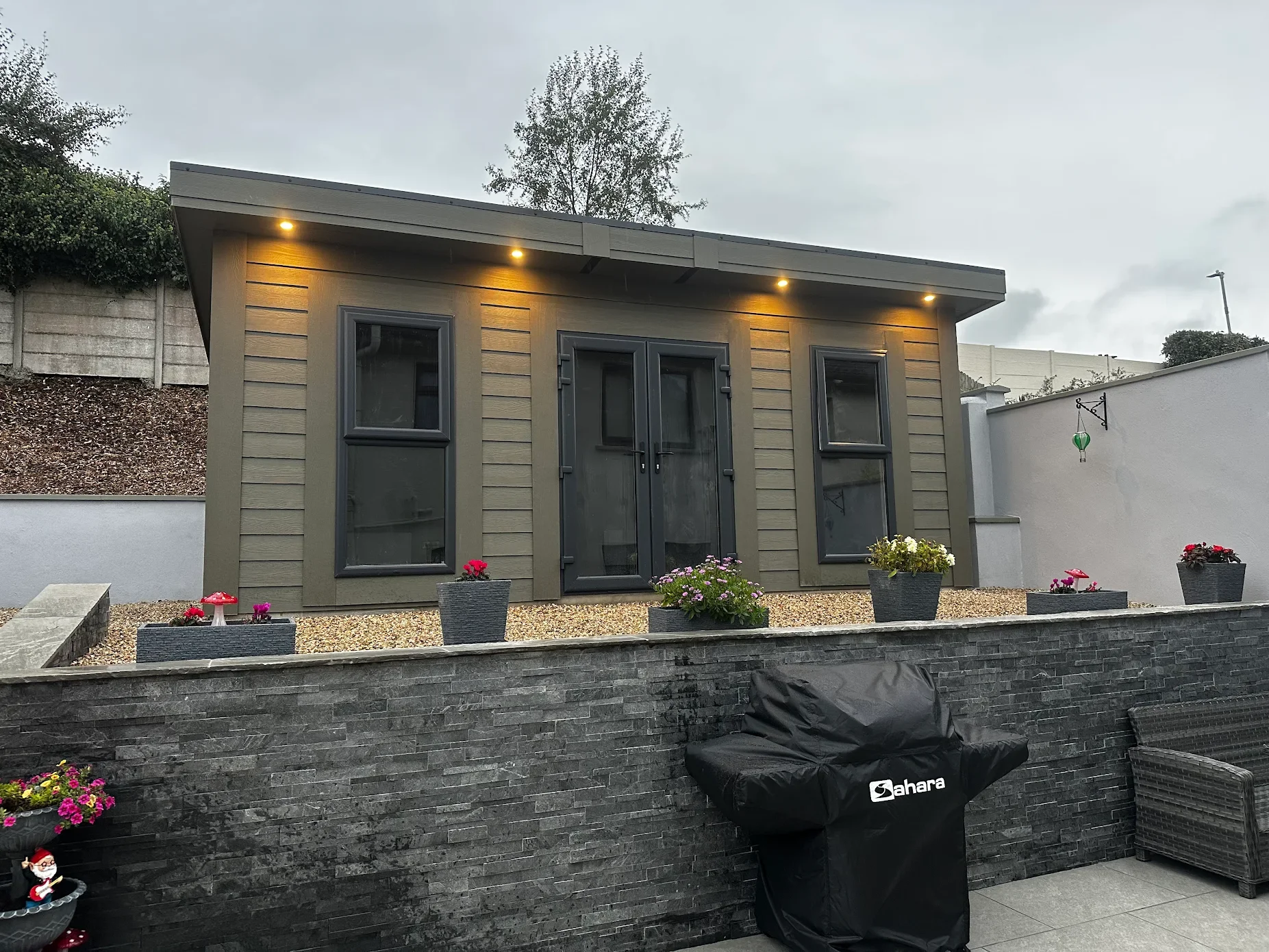 A small gray house with two windows and a glass door, illuminated by four porch lights, is situated behind a stone wall. There are flower pots with pink, red, and white flowers on the wall and a covered barbecue grill in front.