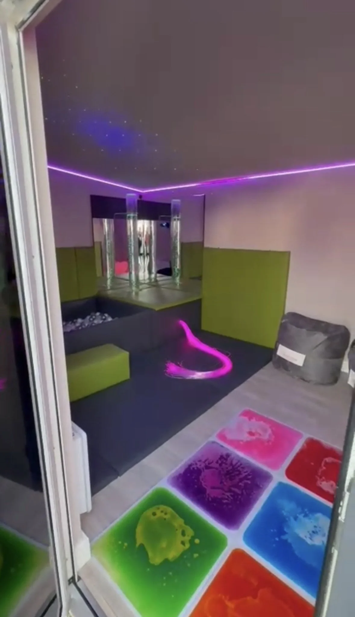 A neon-lit room with glowing colorful floor tiles in green, purple, pink, red, and blue. The ceiling has star-like points of light and purple LED strip lighting. There are lime green wall panels and a cushioned armchair with a pillow in the room.