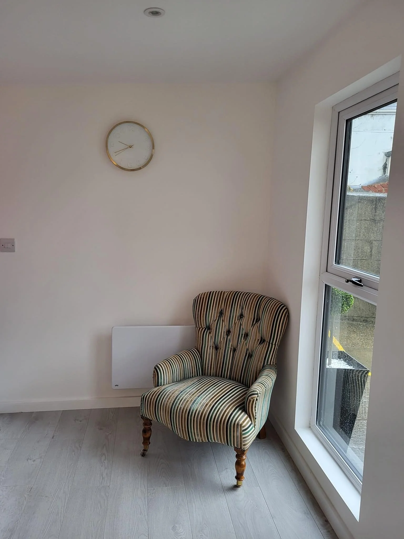 A striped upholstered armchair with wooden legs placed next to a large window in a room with white walls and a light-colored floor. A round wall clock is mounted on the wall above the chair.