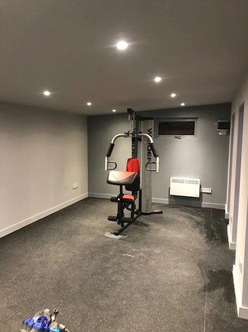 A home gym with a weight machine in a small, minimally decorated room with gray walls and ceiling, carpeted floor, recessed ceiling lights, a small window, and a white wall heater.