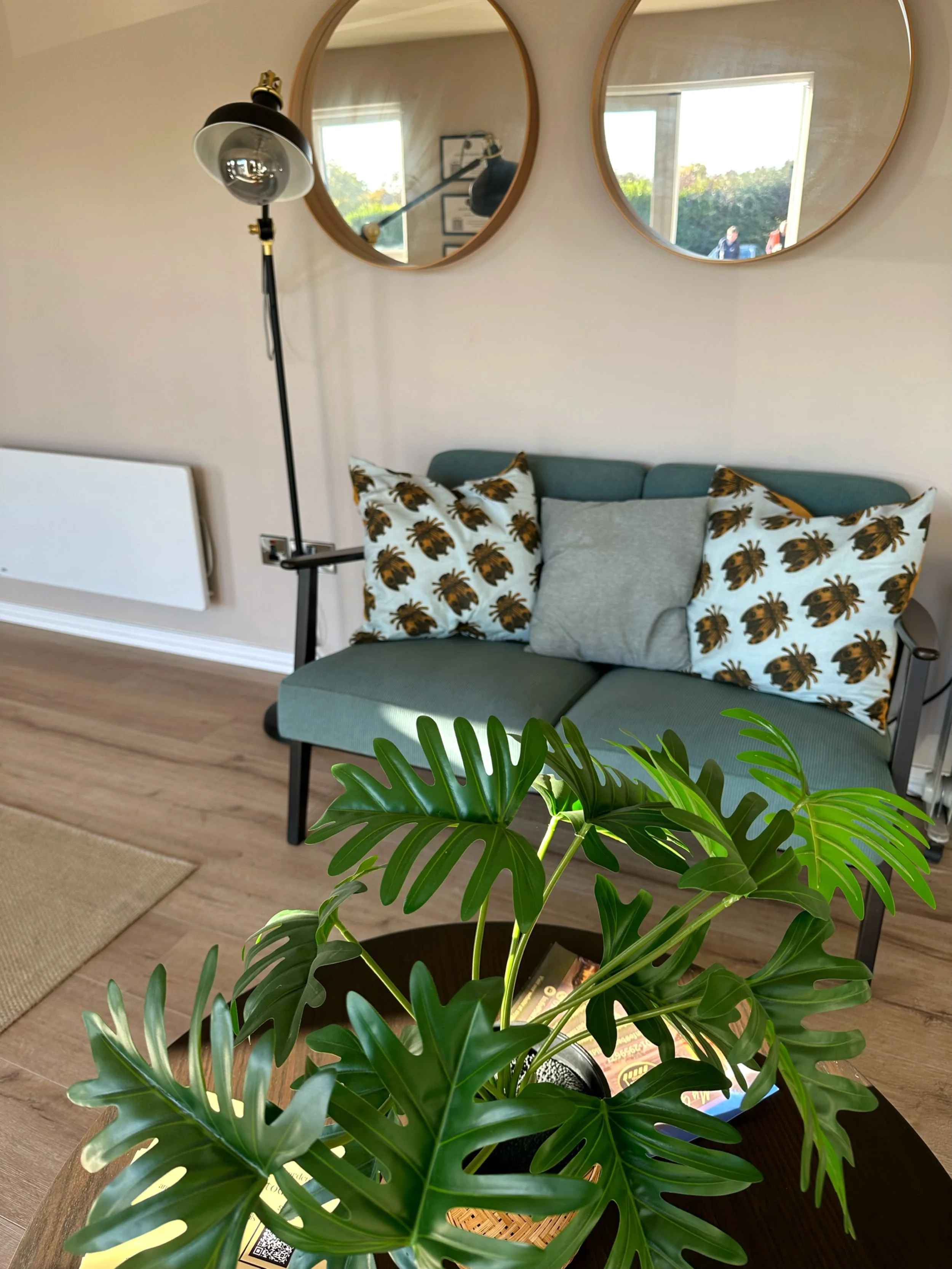 Living room with a plant in the foreground, a teal sofa with pillows, a wooden round coffee table, a standing lamp, two round mirrors on the wall, and sunlight coming through the windows.