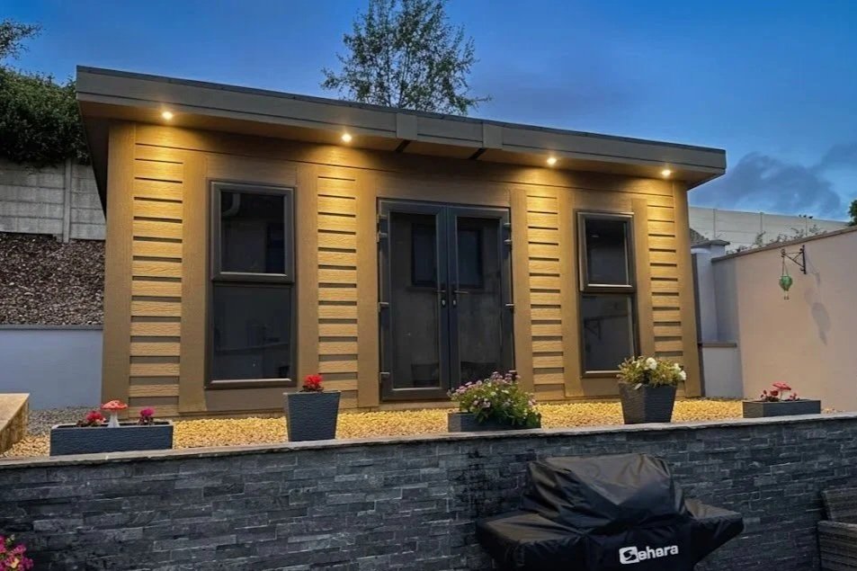 A small modern backyard shed with wood siding, four windows, and glass double doors, illuminated by exterior lights at dusk. In front, potted plants with flowers sit on a gravel bed, and a black barbecue grill cover is visible at the bottom right.