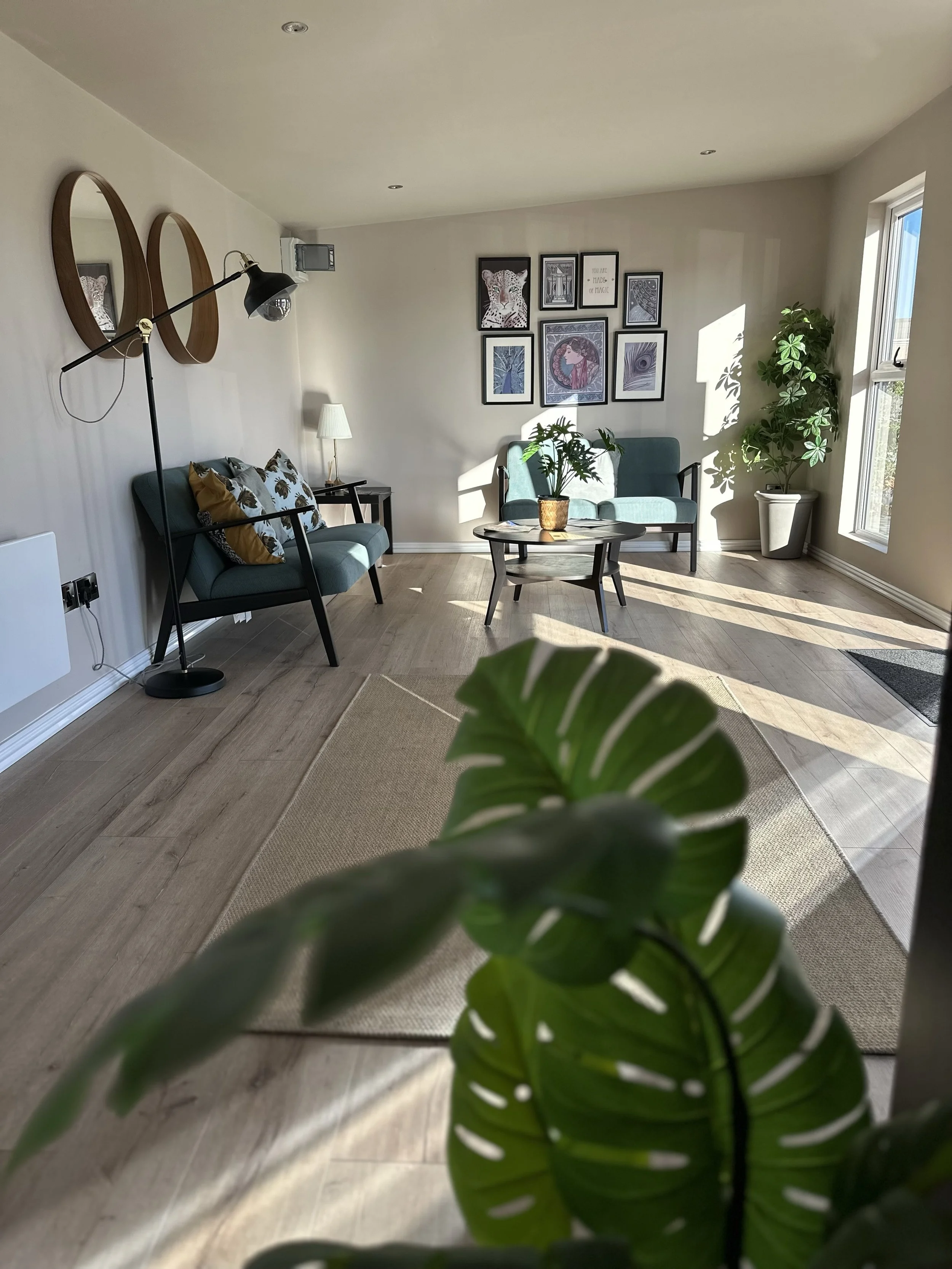 Bright living room with light wood flooring, beige walls, and large window. Two teal sofas, a black coffee table with a potted plant, a side table with a white lamp, and framed art on the wall. Potted plant on the right near the window and a large le