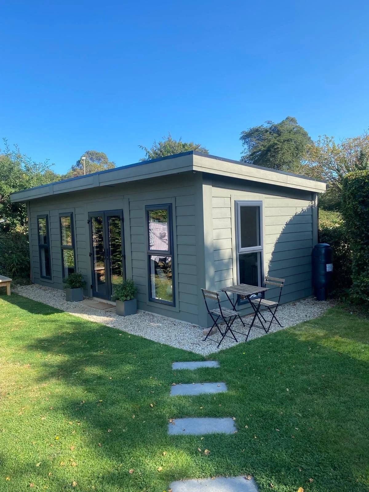 A small, modern, grey building with large windows and a glass door, situated on a lawn with a gravel border, two potted plants outside, and a small outdoor table with two chairs. The background shows trees and a clear blue sky.