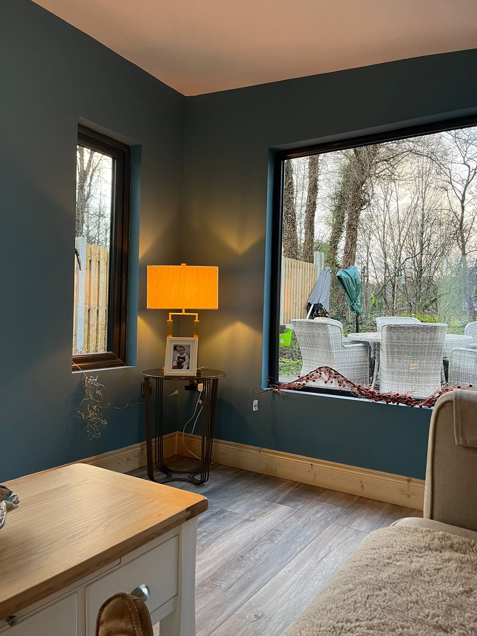 Living room corner with teal walls, a side table with a yellow lamp and framed photo, large window with outdoor patio furniture and trees visible.