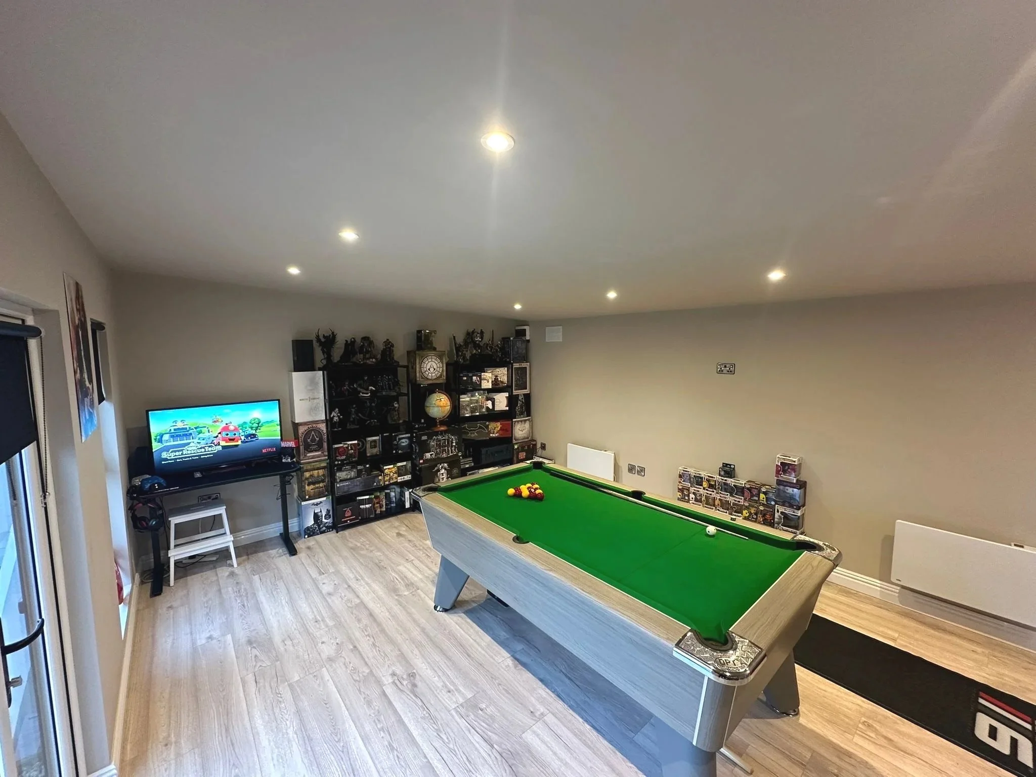 A game room with a pool table, a TV showing animated characters, black shelving filled with collectibles, and a wine rack. The room has light-colored wooden floors and white walls.