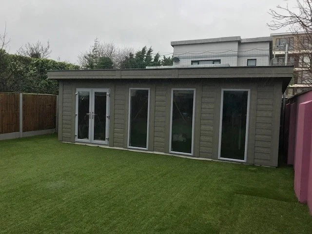 Small backyard shed with large glass doors and windows, situated on a green lawn, with a wooden fence on the left and a purple structure on the right.