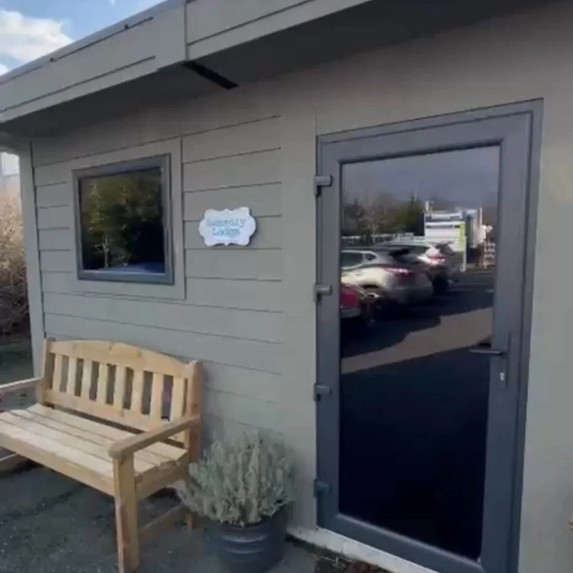 Exterior of a small building with gray siding, a window, and a glass door. There is a wooden bench and a potted plant outside, and a sign that reads 'Laundry' is attached to the wall.