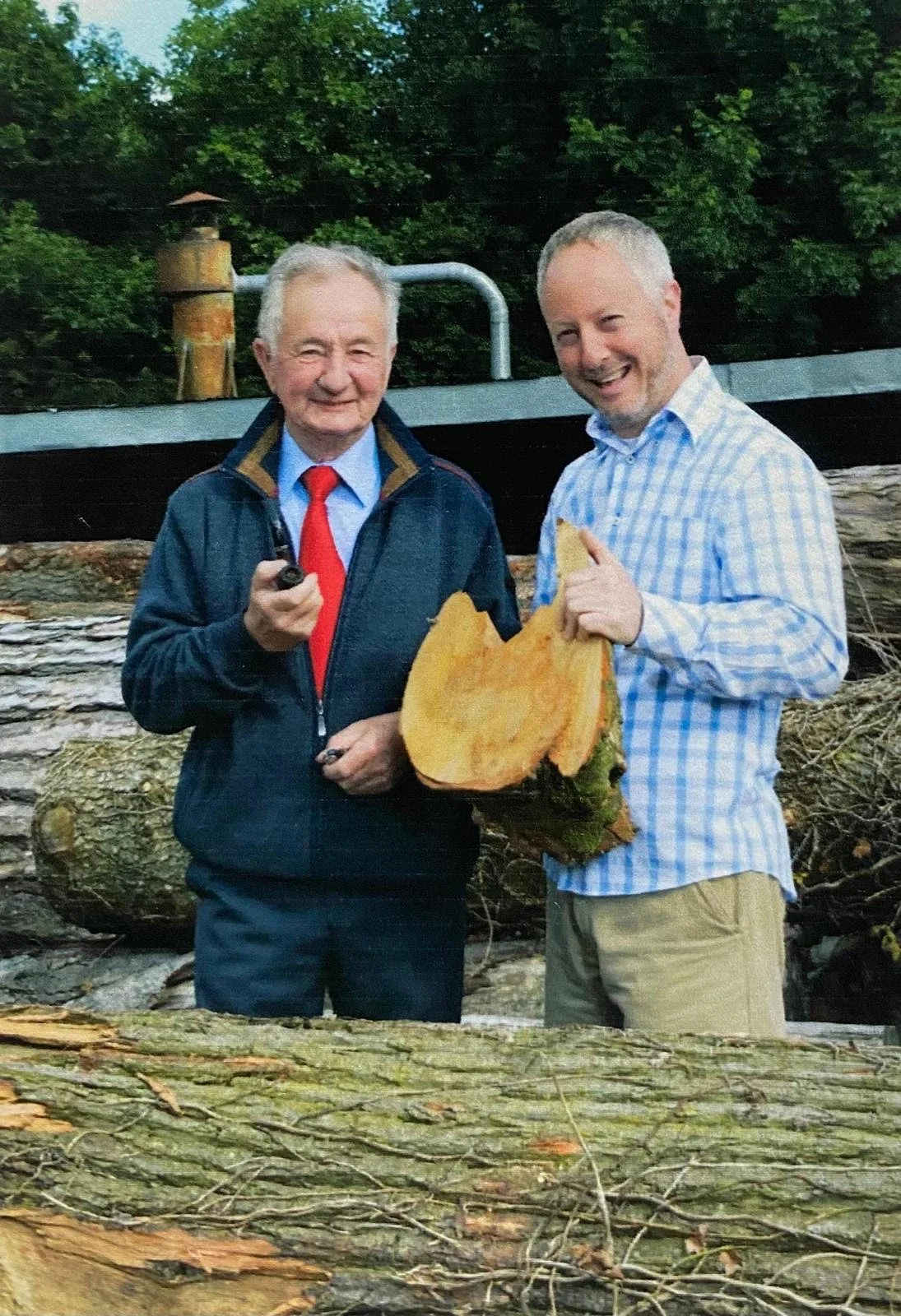 Two men smiling outdoors, one in a suit and red tie holding a pipe, the other in a checkered shirt holding a large piece of a tree trunk, with logs and trees in the background.