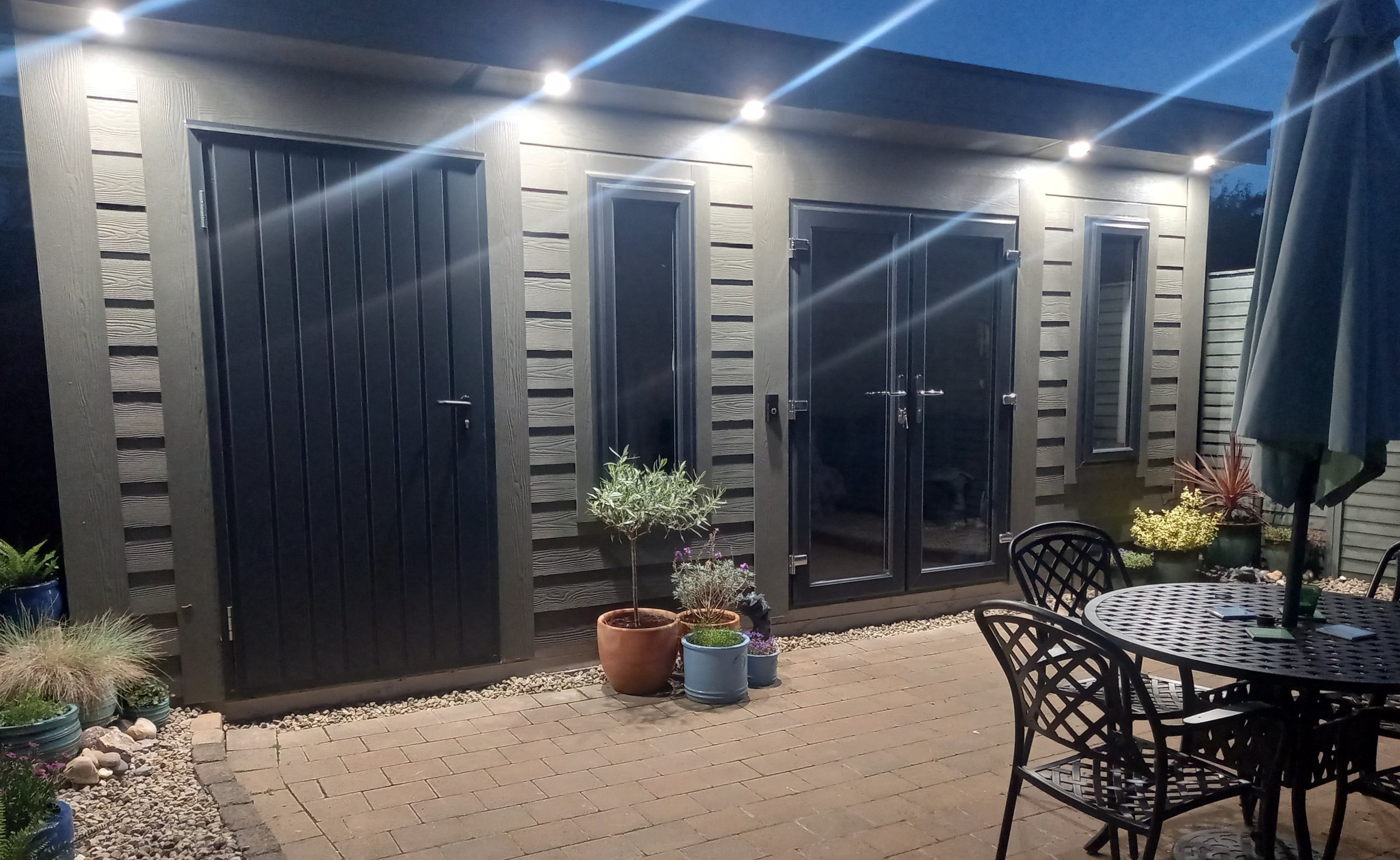 Nighttime view of a backyard patio with outdoor furniture, potted plants, and a deck shed with lights overhead.