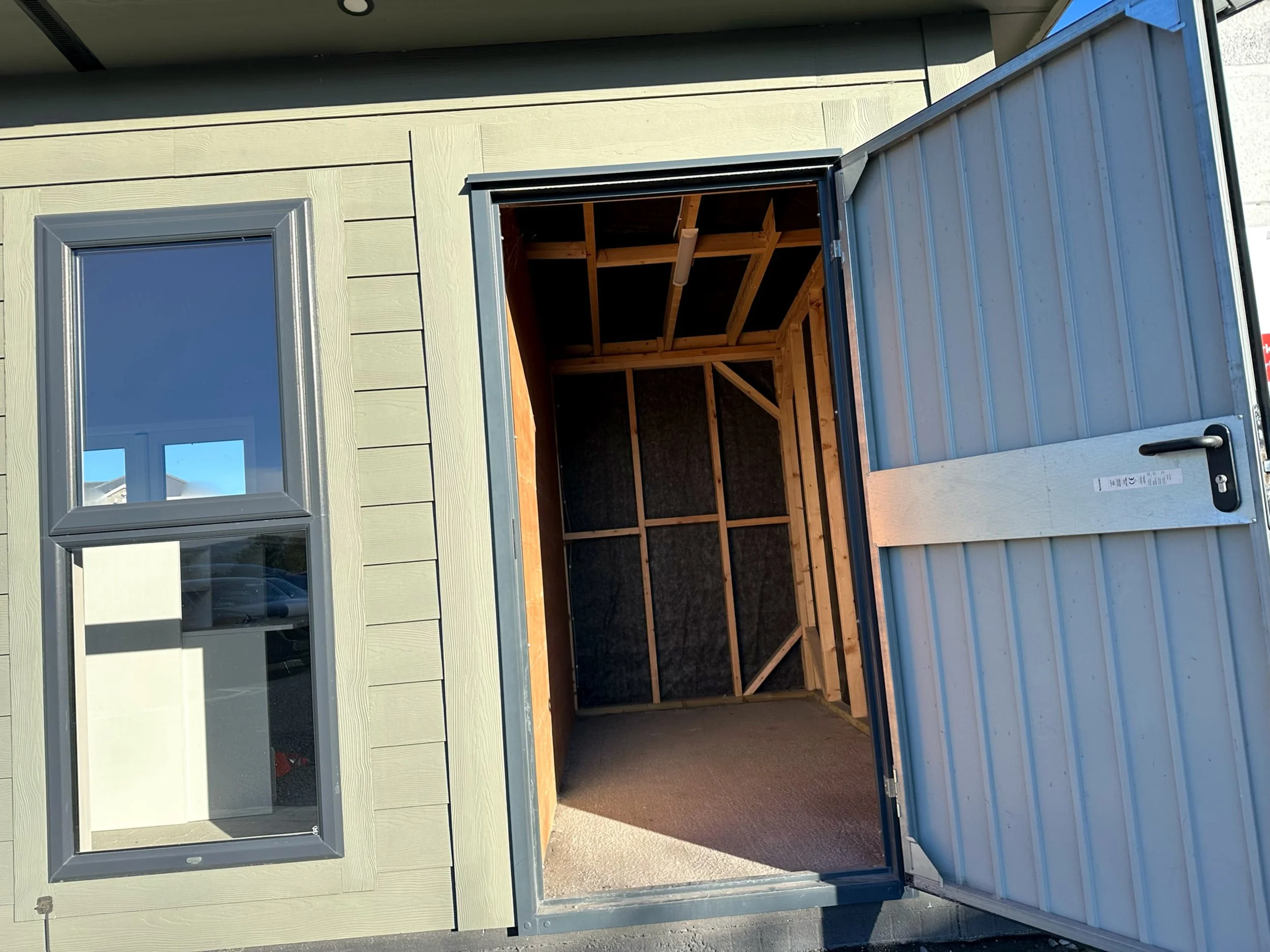 Empty storage shed with partially open metal sliding door, showing wooden framing and interior space, in front of a light-colored house with a window.