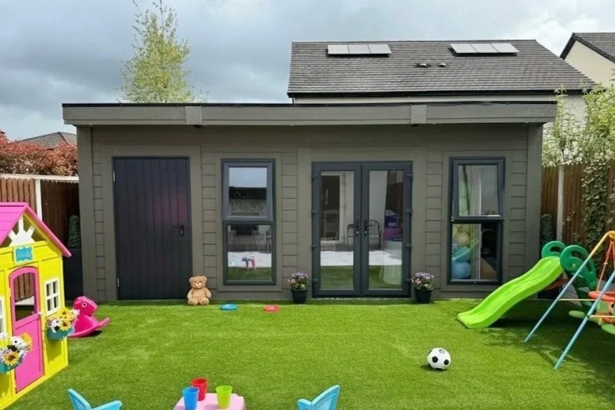 Backyard scene with a gray playhouse, a children's slide, a soccer ball, a plastic playhouse, and children's toys on green artificial grass, with a wooden fence and houses in the background.