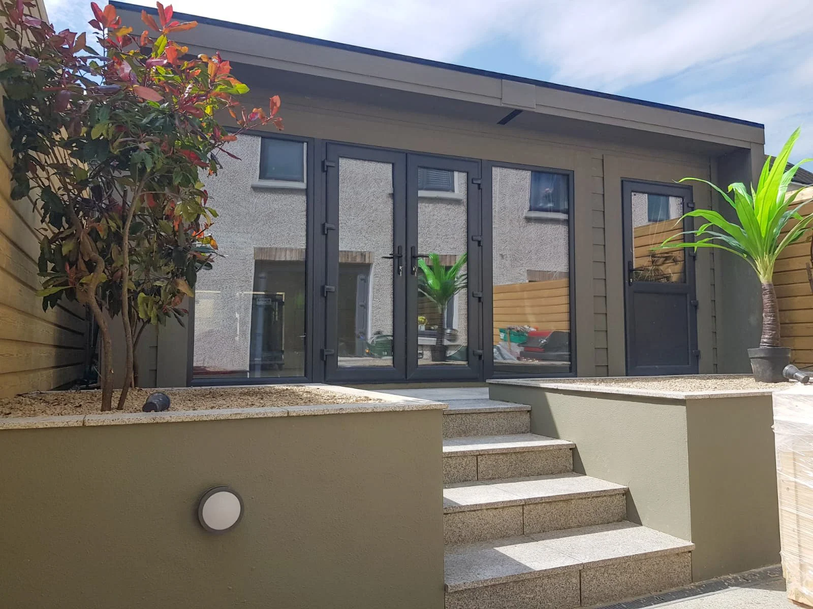 Backyard patio with stairs leading up to a modern glass extension, potted plants, and wooden fences.