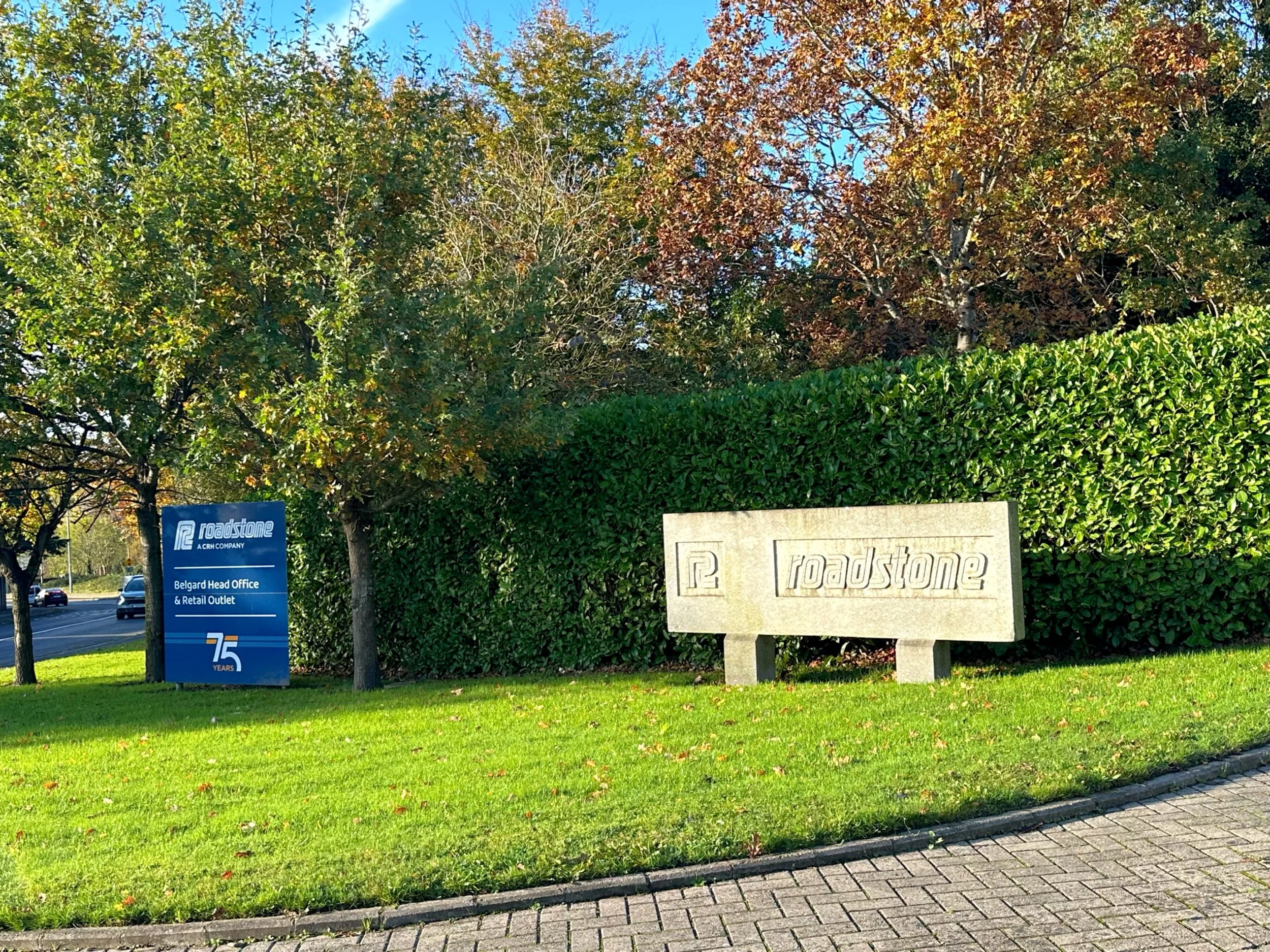 Signage for Roadstone at Belgard Head Office and Retail Outlet, with a concrete sign displaying the Roadstone logo, situated on a grassy area with trees and a hedge in the background.
