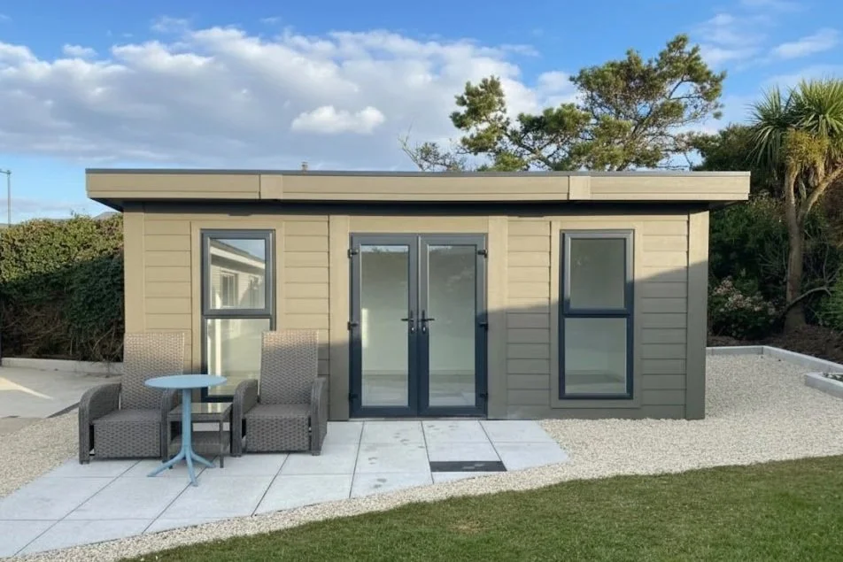 Small modern gray building with glass doors and windows, outdoor seating with two chairs and a small table, surrounded by a landscaped yard and trees under a partly cloudy sky.