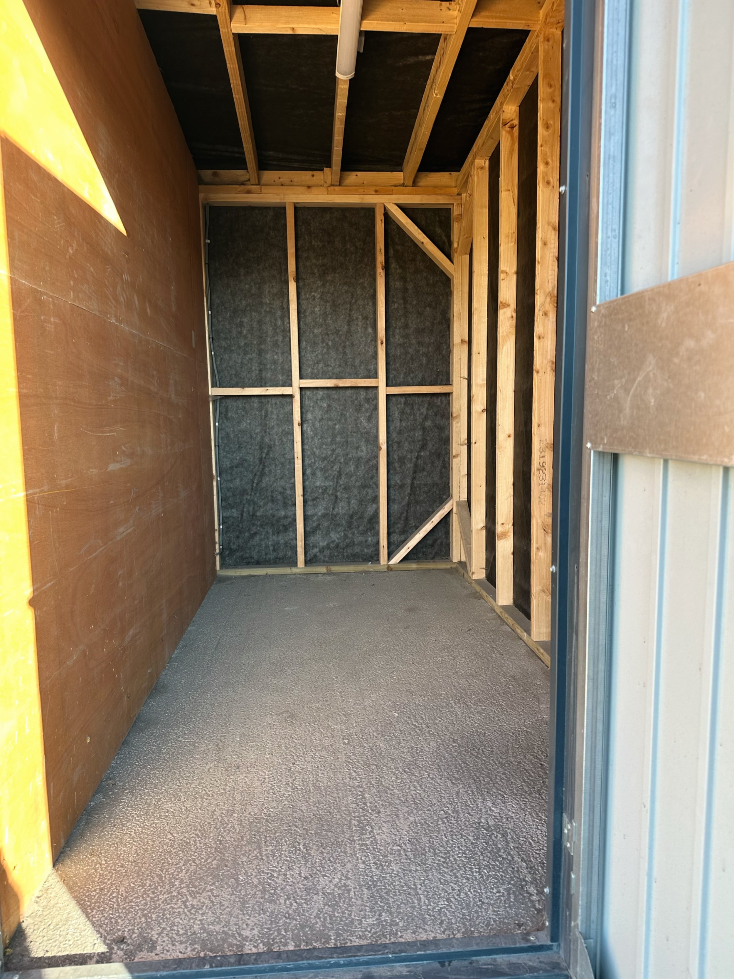Empty room with wooden framing on the walls and ceiling, black insulation or material behind the framing, and a concrete floor.