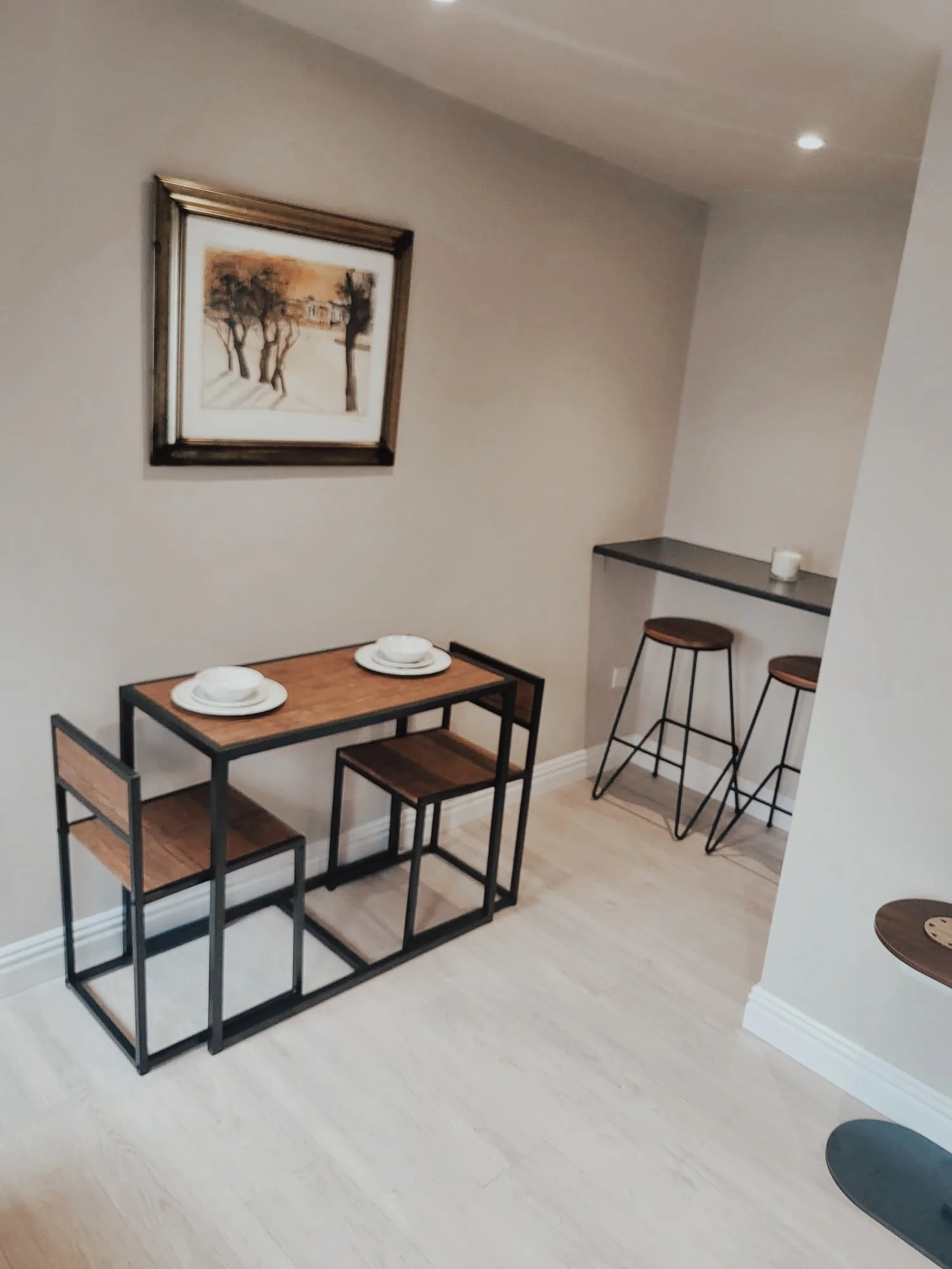 A small dining area with a wooden table set with two white plates and bowls, three matching wooden stools, and a wall-mounted shelf with two additional stools, a candle, and a framed artwork of trees and a building on the wall.