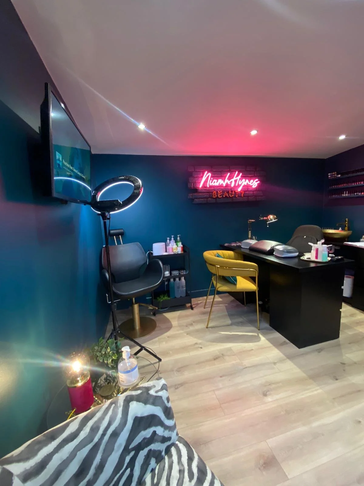 Interior view of a beauty salon with a neon pink sign on a dark brick wall reading 'Niamh Hynes Beauty'. The salon has a black desk with various manicure supplies, a yellow chair, a black salon chair, and a small shelving unit with beauty products. There is a large flat-screen TV on the wall, a ring light, and a bright sofa in the foreground. The floor is light wood and the lighting is a combination of ceiling spotlights and ambient light.