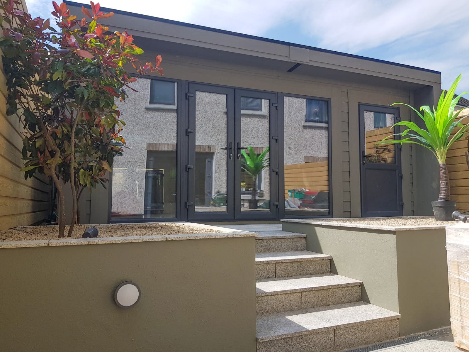 A small building with large glass doors, elevated by stairs, surrounded by plants and wooden fences under a blue sky.