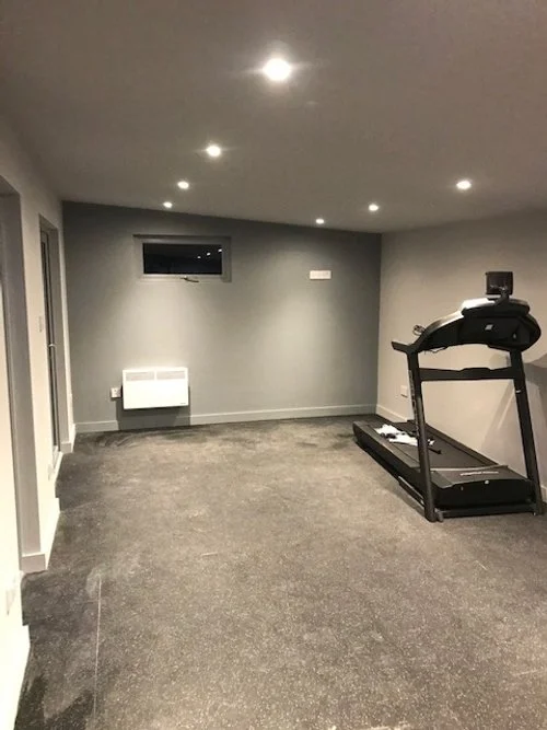 Empty basement room with a treadmill on the right side, gray walls, carpeted floor, and a small window near the ceiling.