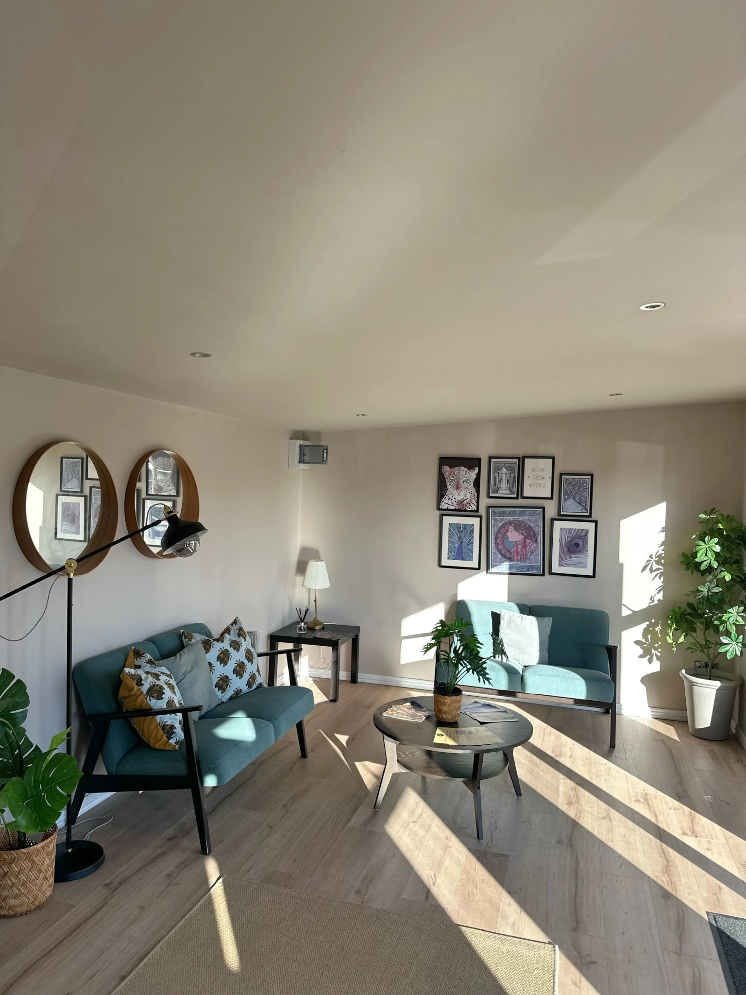 Sunlit living room with two teal sofas, a round coffee table with magazines and a potted plant, framed pictures on the wall, a floor lamp, and potted plants near the window.
