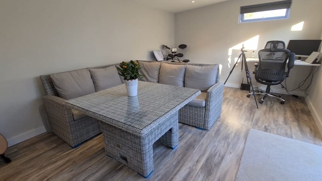 A room with a sectional wicker sofa with beige cushions, a table with a potted plant on it, a window with sunlight, a desk with a computer monitor and office chair, a tripod with a camera, and some books and tablets on the desk.