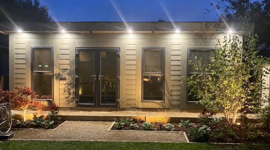 Exterior view of a single-story house at night, featuring three windows and a glass door, with outdoor lighting and landscaped garden plants.