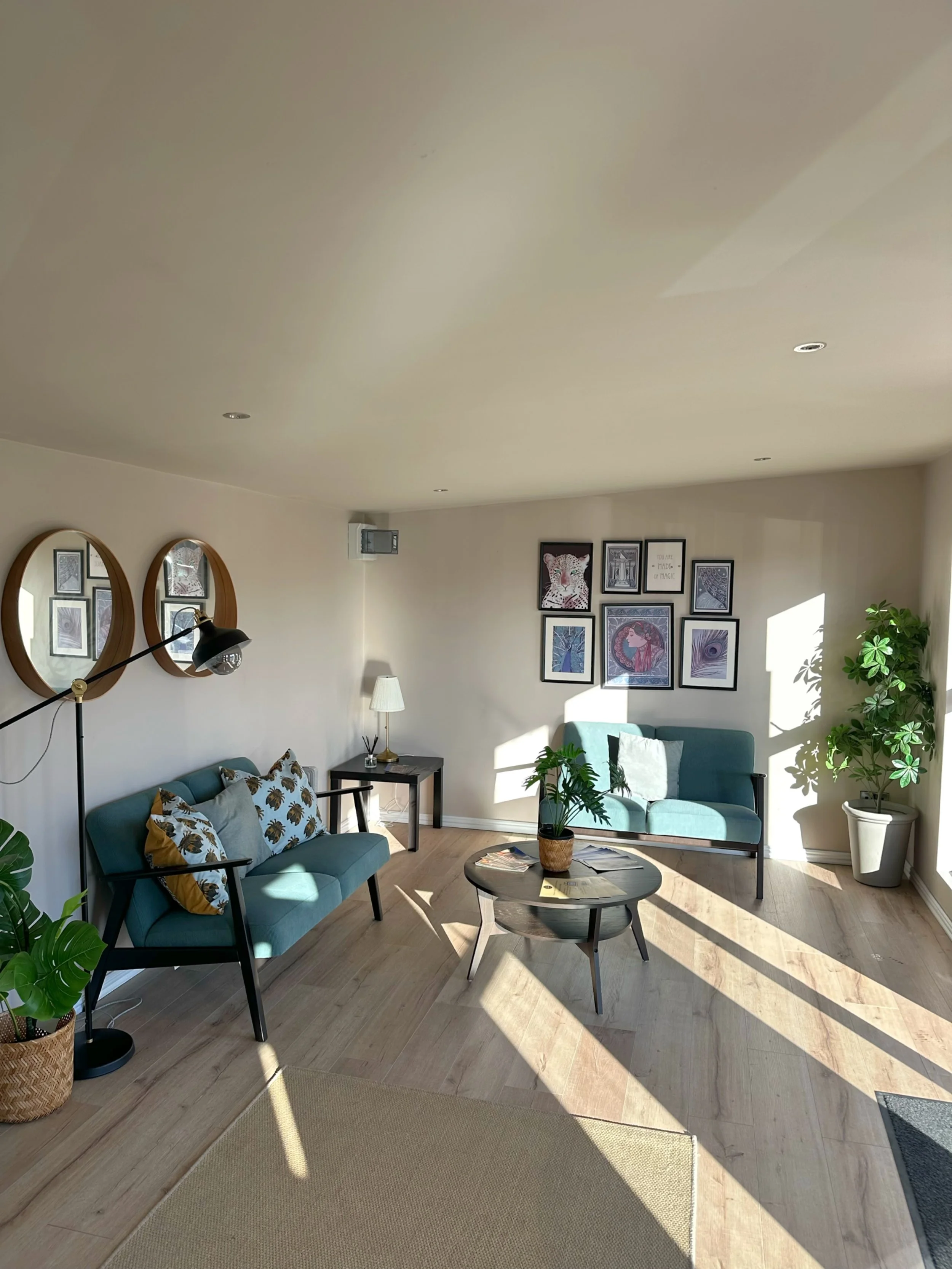Living room with two teal sofas, one larger with pillows, and one small. Coffee table with plants and magazines. Wall gallery with framed art. Potted plants, side table with lamp, wall-mounted mirror, and natural light streaming through windows.