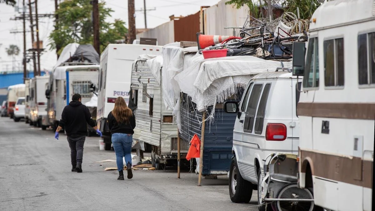 People walking past a lineup of abandoned and damaged RVs and trailers on a street.